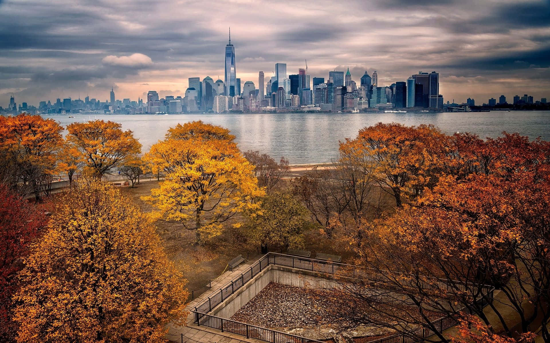 Fall City 桌布 壁紙 — 秋天的樹木帶著火紅的橙色葉子，框住了紐約市天際線在陰沉多雲天空下的景色。