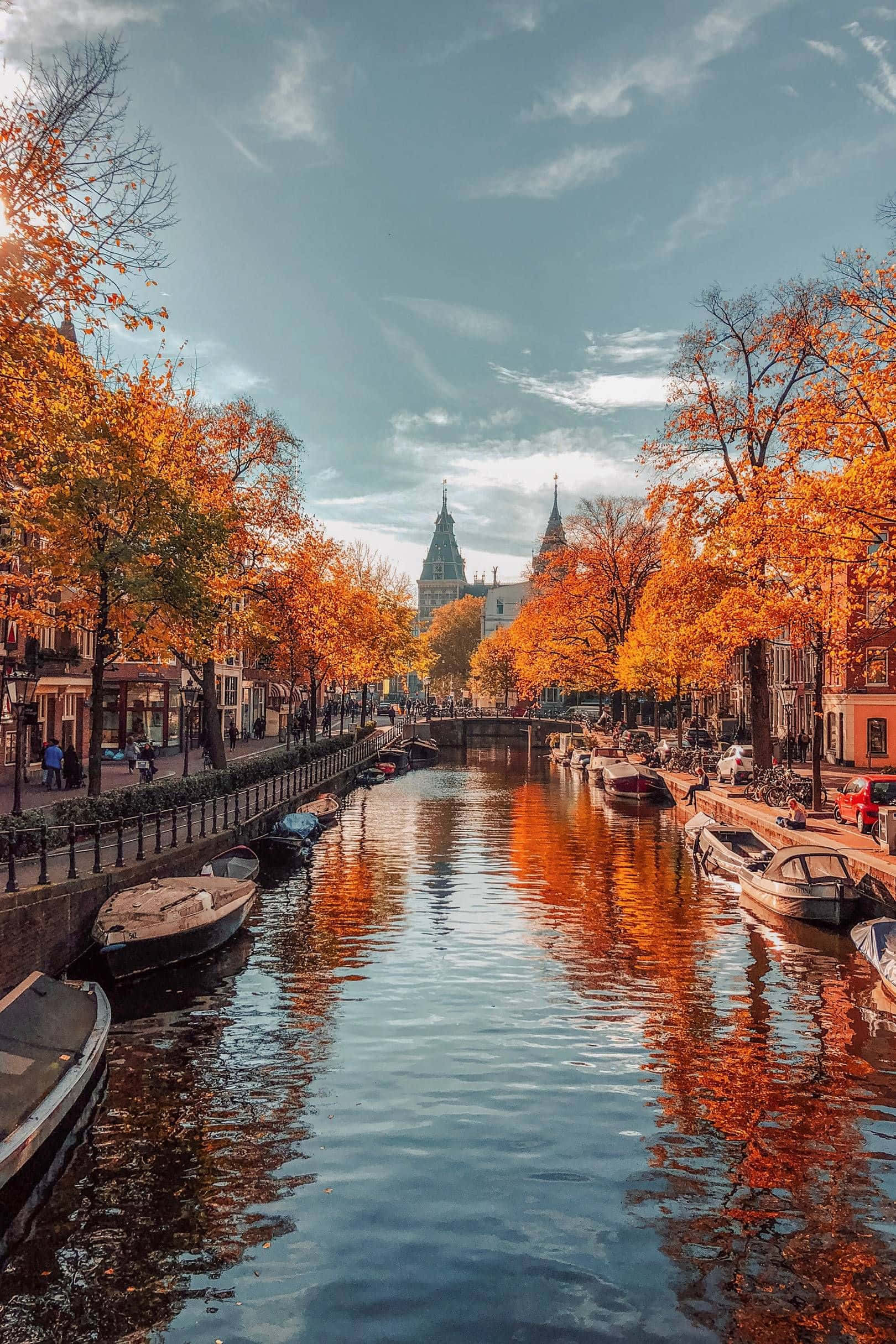 Beautiful Autumn 桌布 壁紙 — 秋天的樹葉在寧靜的運河旁閃耀著橙色，歷史建築和船隻在柔和的藍天下倒映在平靜的水面上。