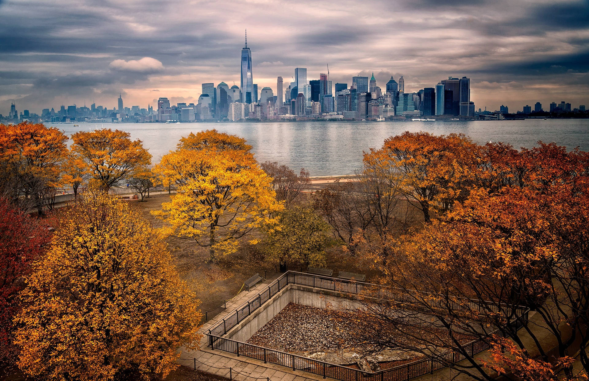 New York City Desktop 桌布 壁紙 — 秋日樹木以火紅橙葉框住紐約市天際線景觀，背景是陰沉多雲的天空。