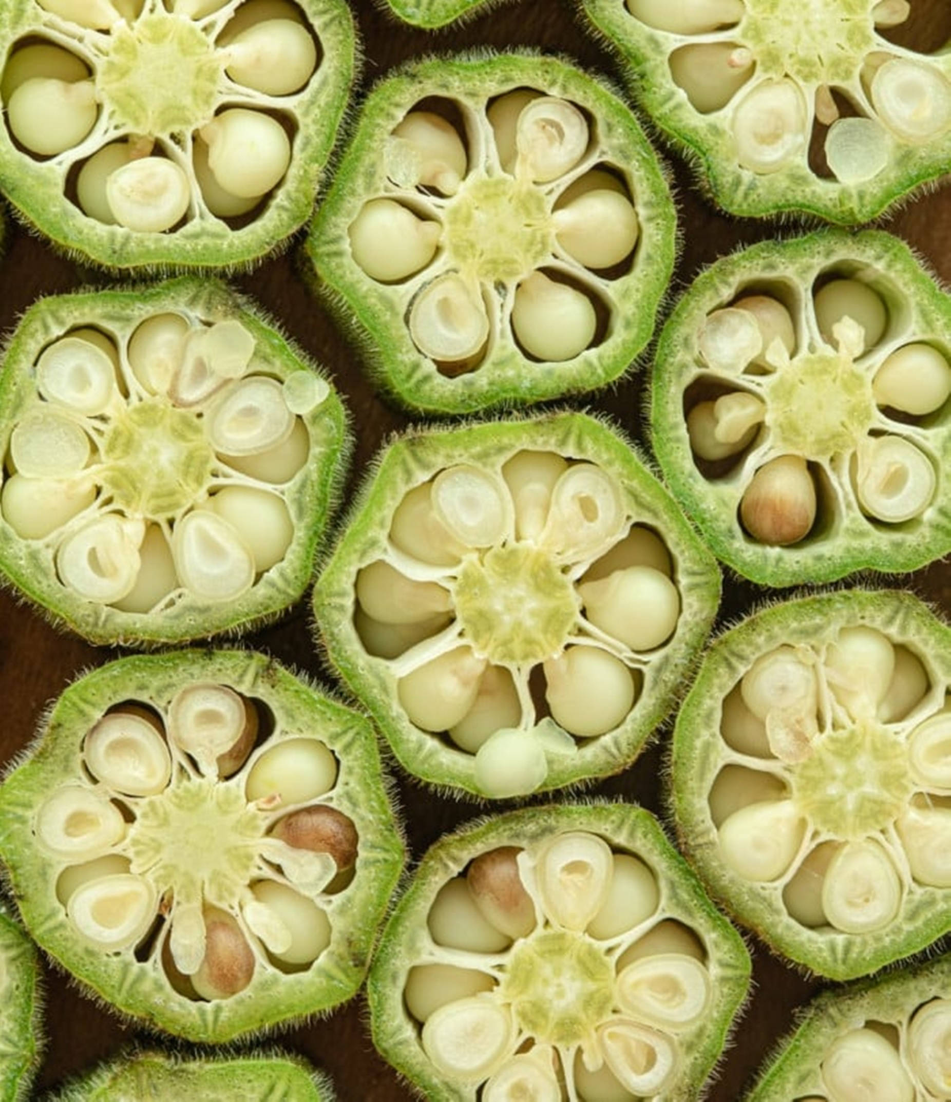 Fruits And Vegetables 桌布 壁紙 — 秋葵莢的橫截面揭示了淡色種子嵌套在綠色腔室中，在深色木質表面上的複雜圖案。