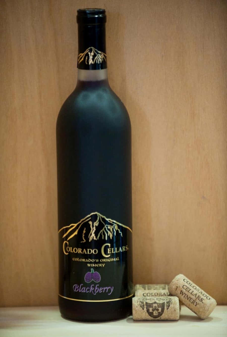 Blackberry Wine 桌布 壁紙 — 科羅拉多酒莊黑莓酒的時尚黑色酒瓶，旁邊放著三個品牌軟木塞，背景為溫暖的木質。