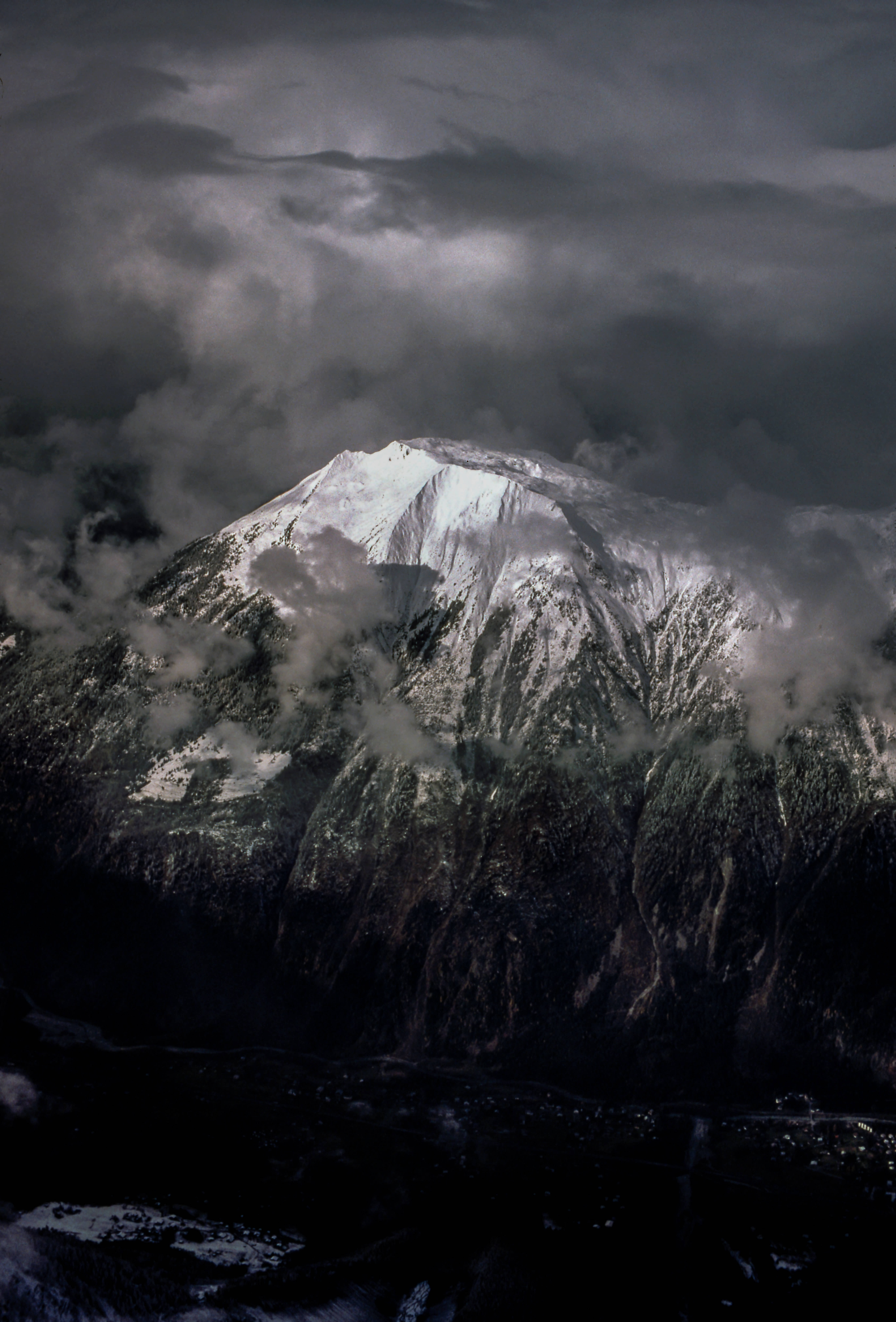 黑色 iPhone 桌布 桌布 壁紙 — 積雪的山峰刺穿黑暗的暴風雲，戲劇性的光線揭示了下方崎嶇的懸崖。