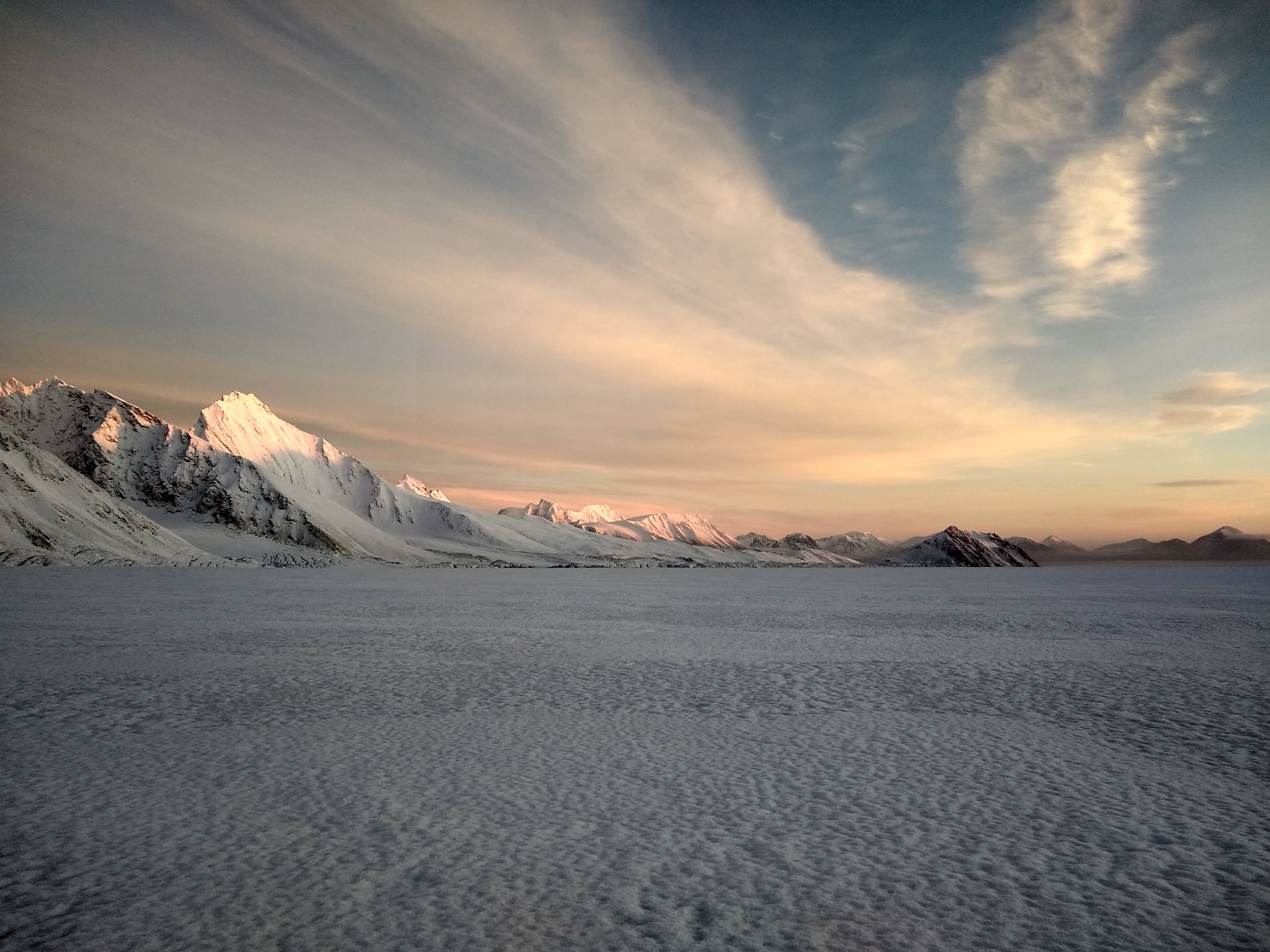 Svalbard 桌布 壁紙 — 積雪的山峰在廣闊冰原上柔和的日落天空下閃耀。