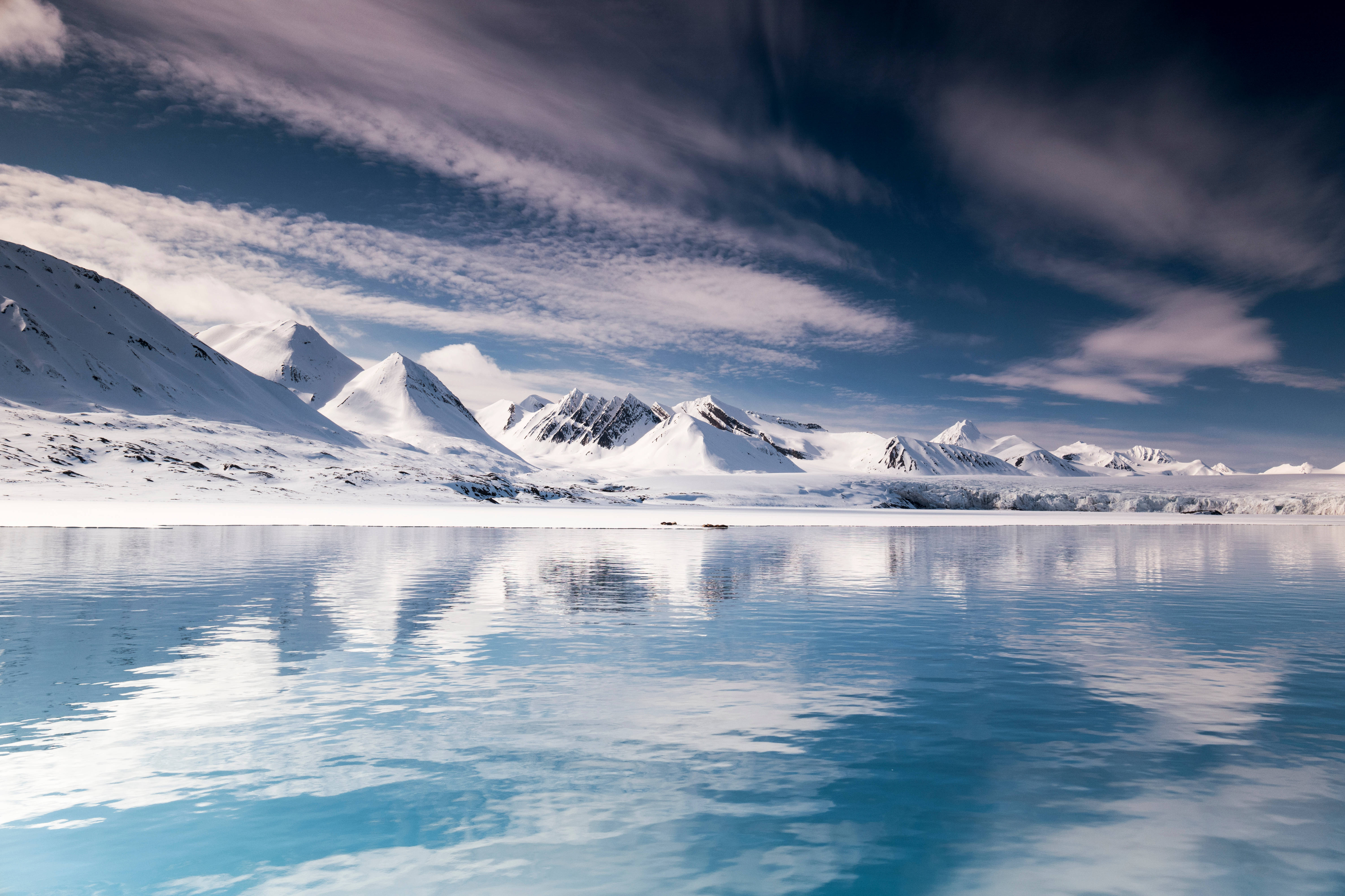 Svalbard 桌布 壁紙 — 積雪的山峰在戲劇性的多雲天空下完美地倒映在玻璃般的藍色湖泊中。