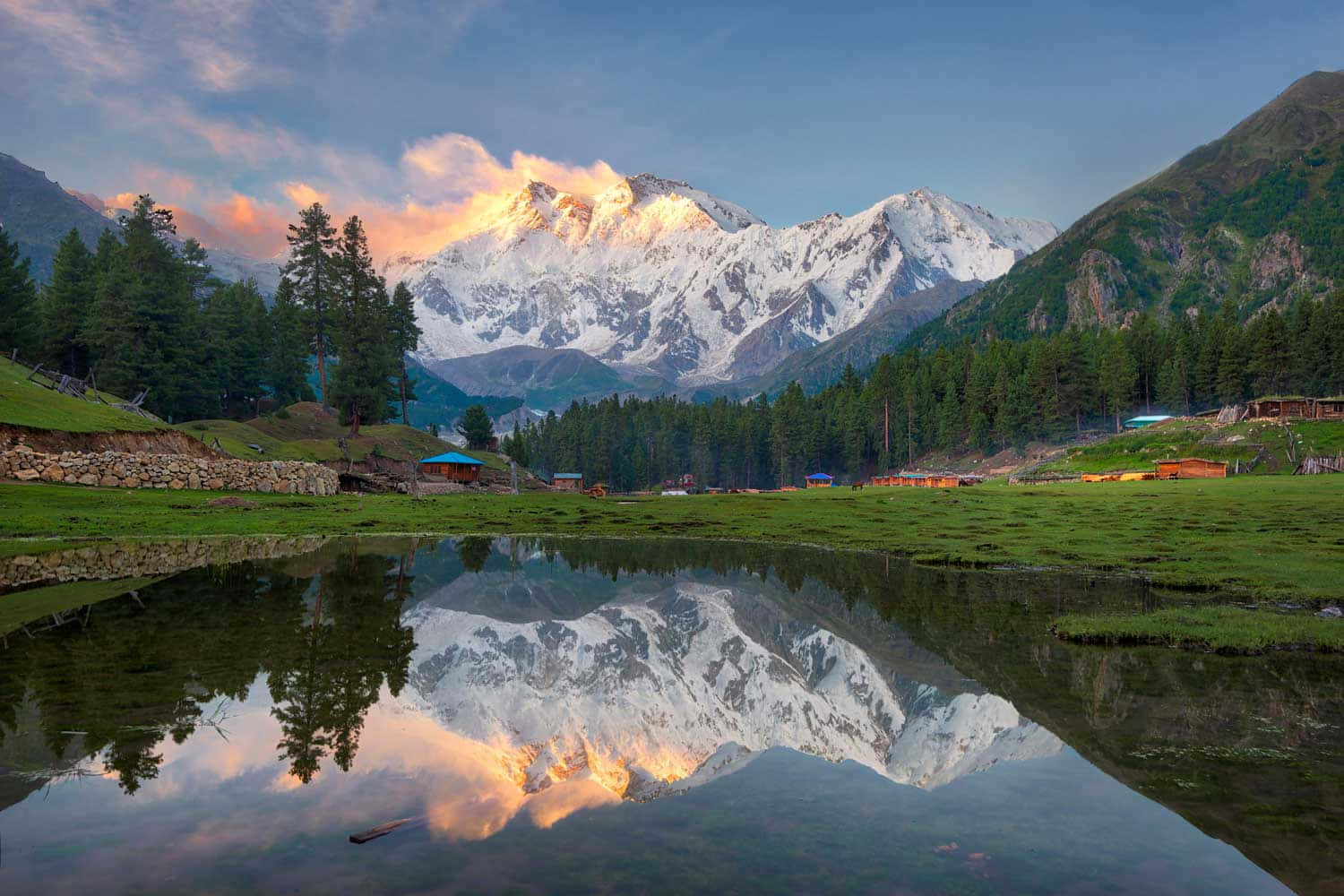 Pakistan 桌布 壁紙 — 積雪的山峰在日出時閃耀，完美倒映在寧靜的高山湖泊中，周圍環繞著松林和質樸的小屋。