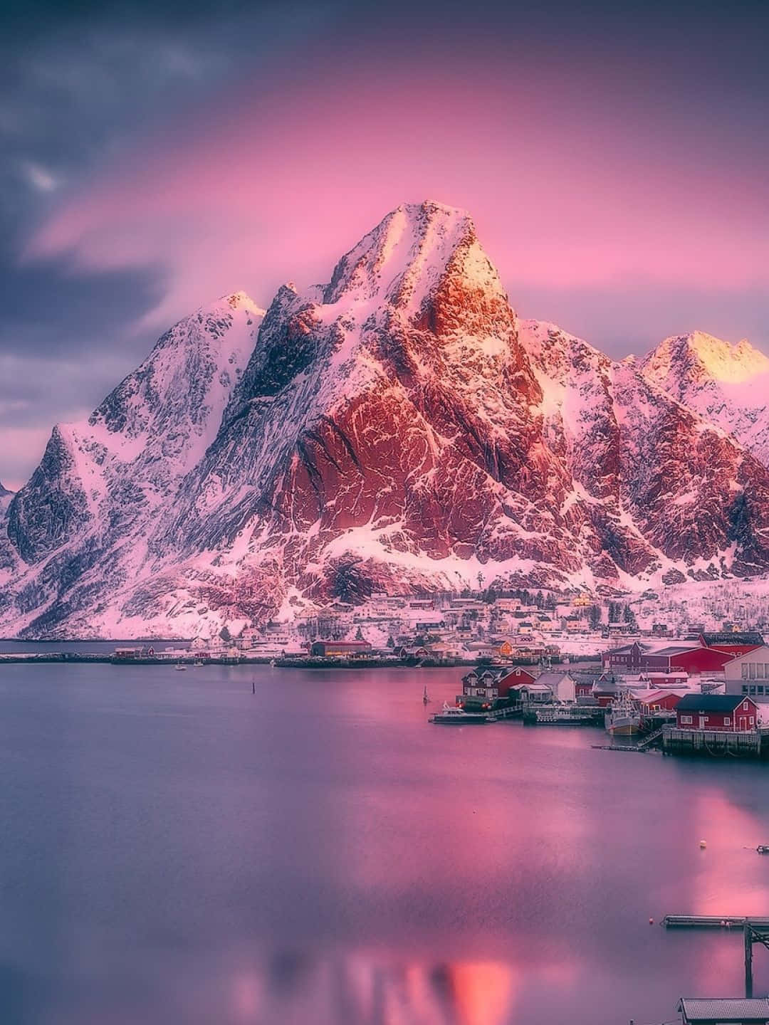 自然 桌布 壁紙 — 積雪的山峰在日落時分泛著粉紅色的光芒，映照在如鏡面般的峽灣上的一個寧靜漁村。