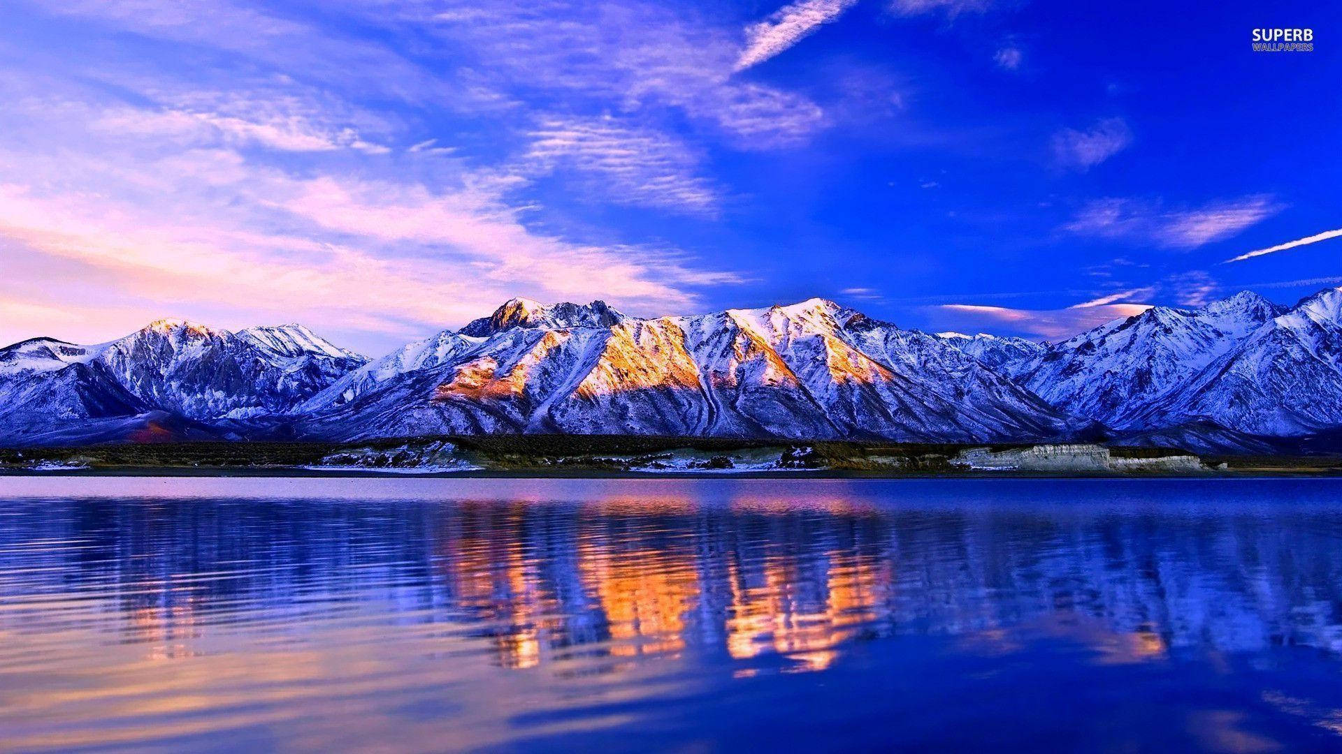 風景 桌布 壁紙 — 積雪的山峰在日落時閃耀著金色的光芒，完美地倒映在平靜的藍色湖面上，天空色彩斑斕。