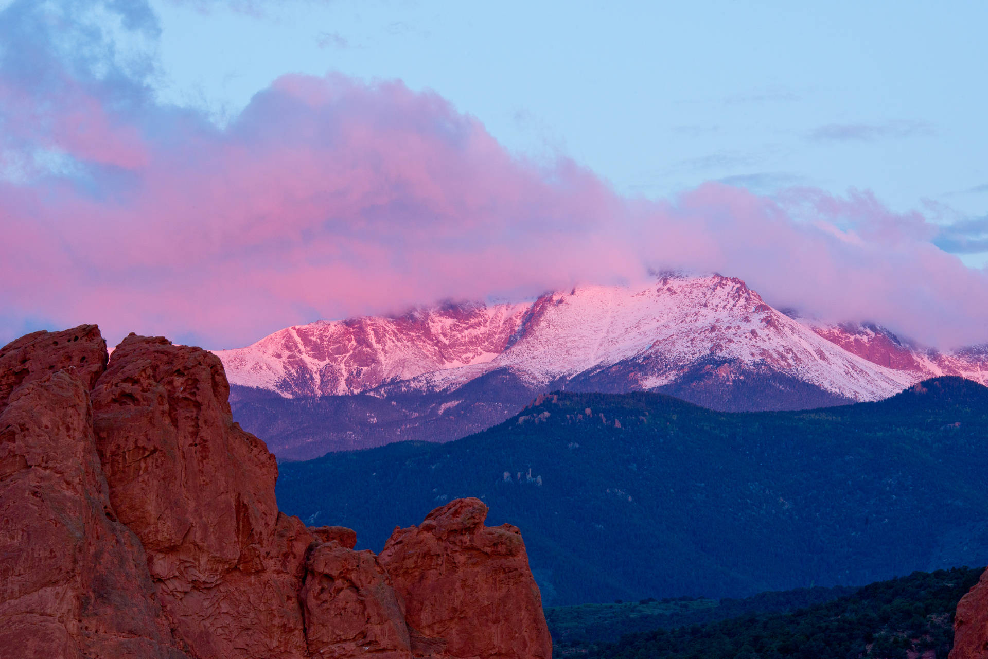 Colorado 桌布 壁紙 — 積雪的山峰在暮色雲層下呈現粉紅色光芒，前景有紅色岩石 formations。