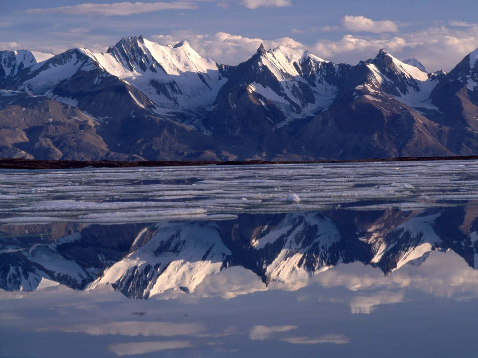 Greenland 桌布 壁紙 — 積雪的山峰在柔和的藍天和散落的雲朵下，完美地倒映在結冰的湖面上。