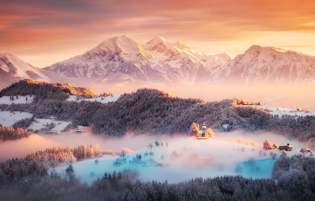 Slovenia 桌布 壁紙 — 積雪的山峰在溫暖的日出光芒中閃耀，薄霧籠罩著森林覆蓋的山谷，山谷中的小山上有一座小教堂。