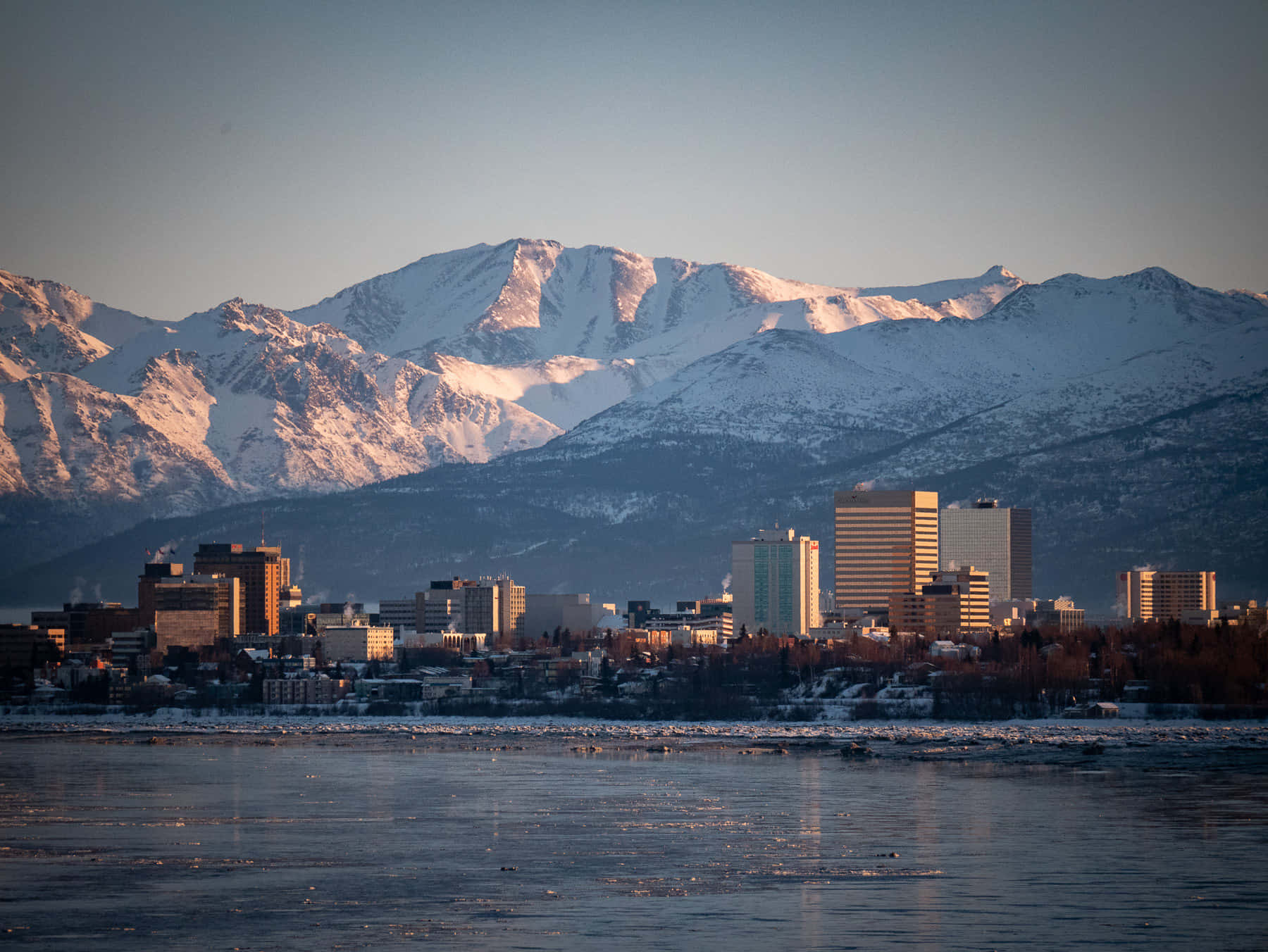 Mountain Landscape 桌布 壁紙 — 積雪的山峰在金色日出光芒中閃耀，背後是城市天際線倒映在部分結冰的河流中。