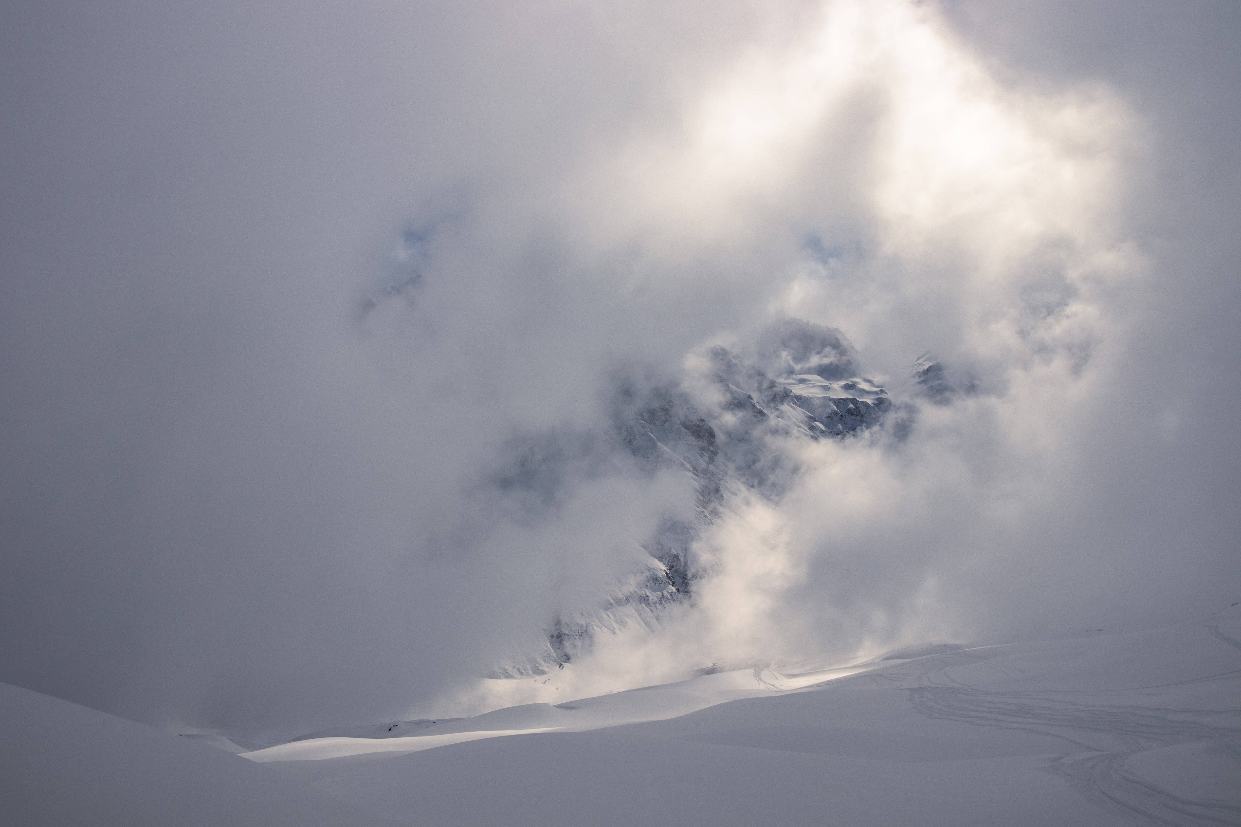 白雪公主 桌布 壁紙 — 積雪的山峰從旋轉的雲層中浮現，陽光穿透雲層照射在帶有滑雪痕跡的雪原上。