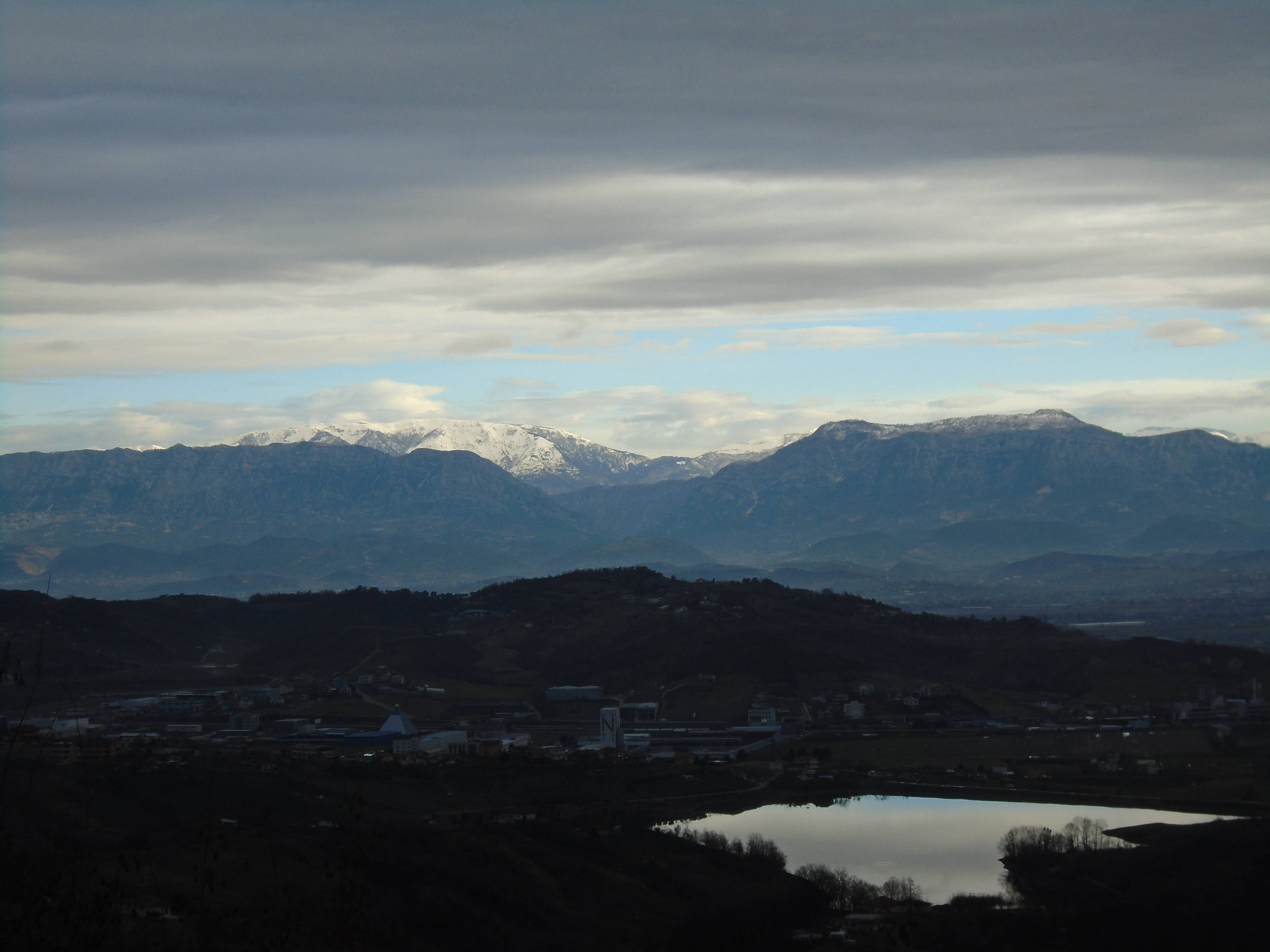 Albania 桌布 壁紙 — 積雪的山峰聳立在山谷之上，山谷中有平靜的湖泊和遠處的小鎮，天空多雲。