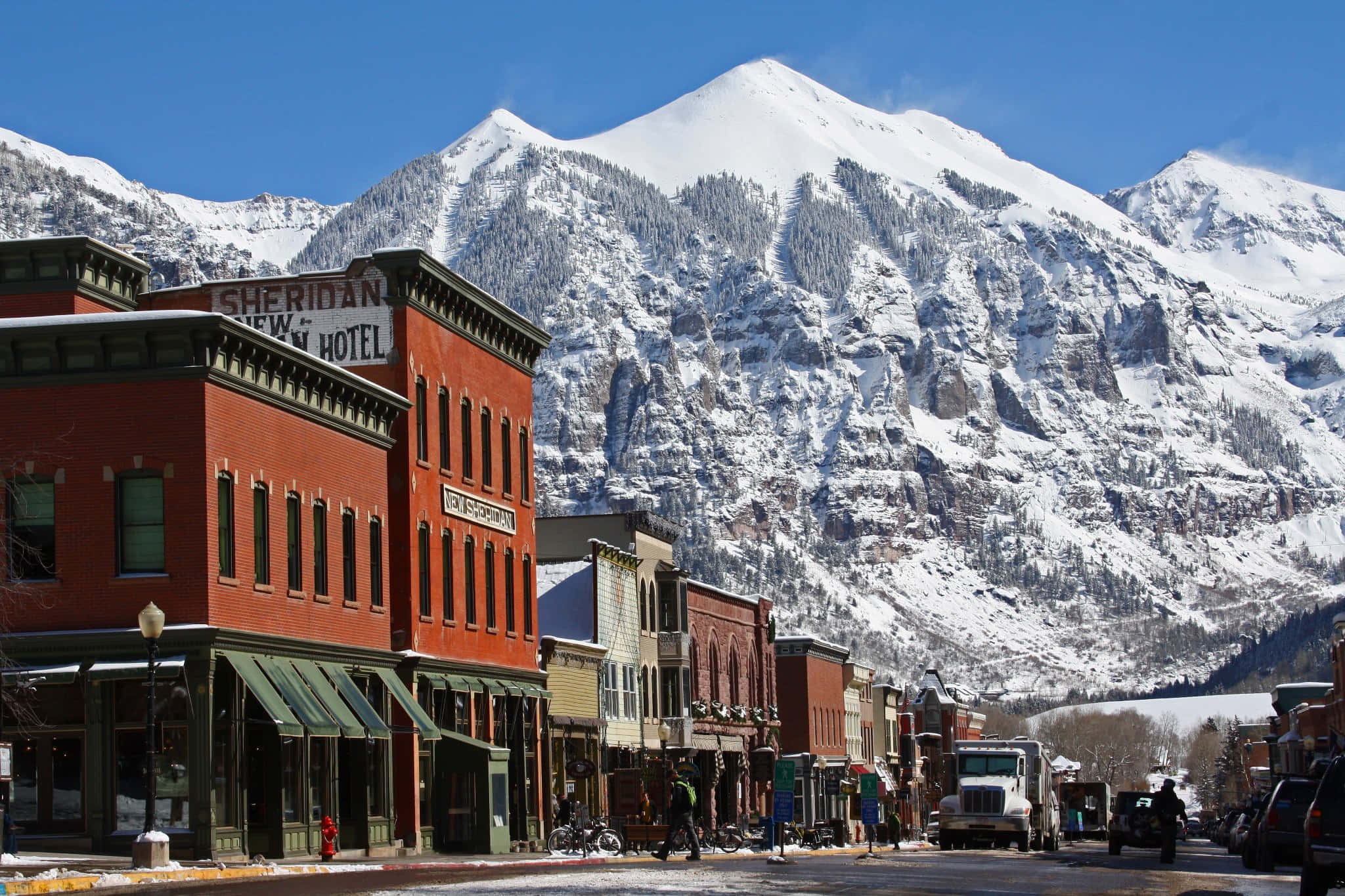 Colorado 桌布 壁紙 — 積雪的山峰聳立在歷史悠久的紅磚小鎮街道後方，街道上有復古商店門面和清澈的藍天。