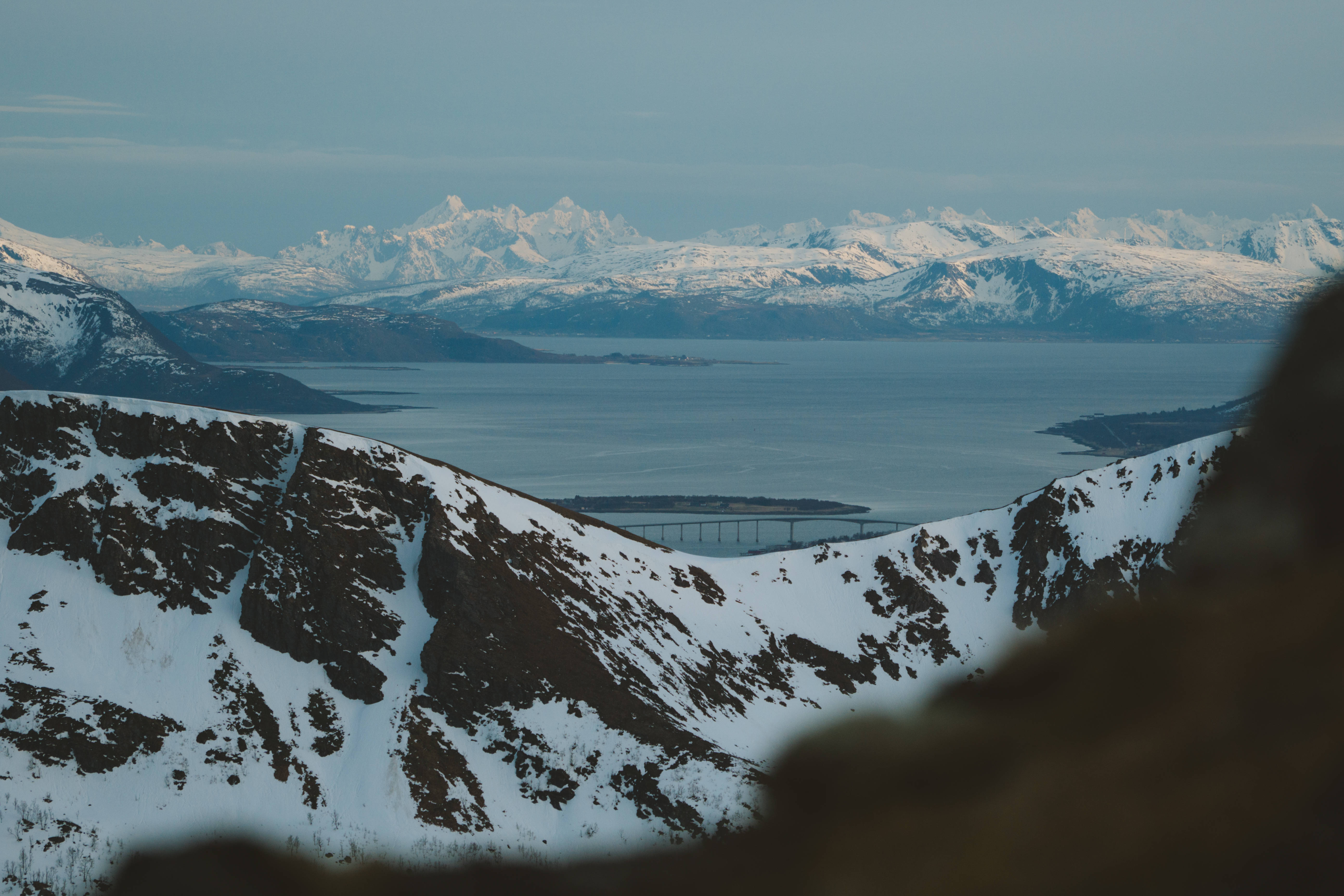 冬季桌面 桌布 壁紙 — 積雪的山脈框住了一個廣闊的峽灣，遠處有一座橋樑，天空柔和。