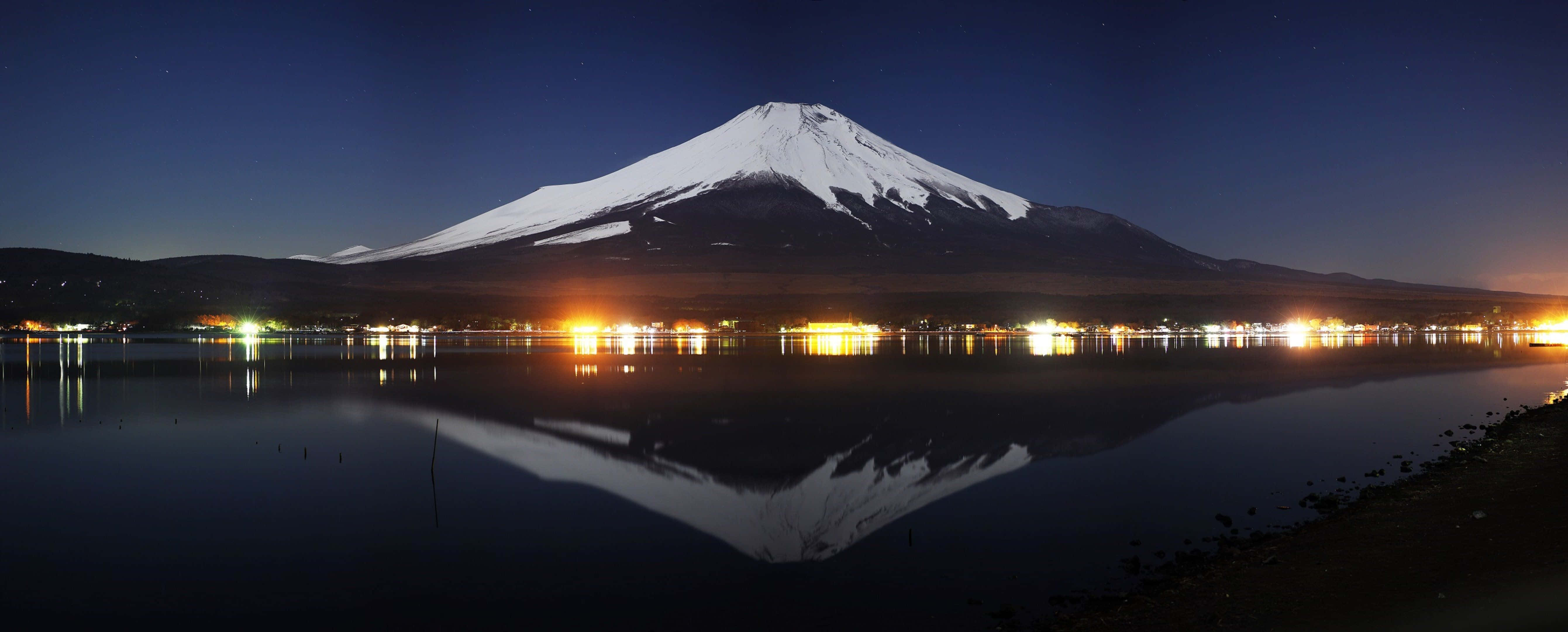 日本 桌布 壁紙 — 積雪覆蓋的富士山在星空下平靜的湖面上完美倒映，岸邊燈光閃爍。