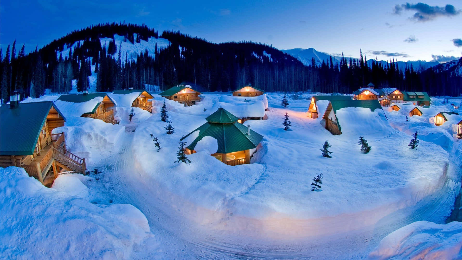 Christmas Winter Wonderland 桌布 壁紙 — 積雪覆蓋的小屋在深藍色天空下，以暮色山巒為背景，溫暖地閃耀著光芒。