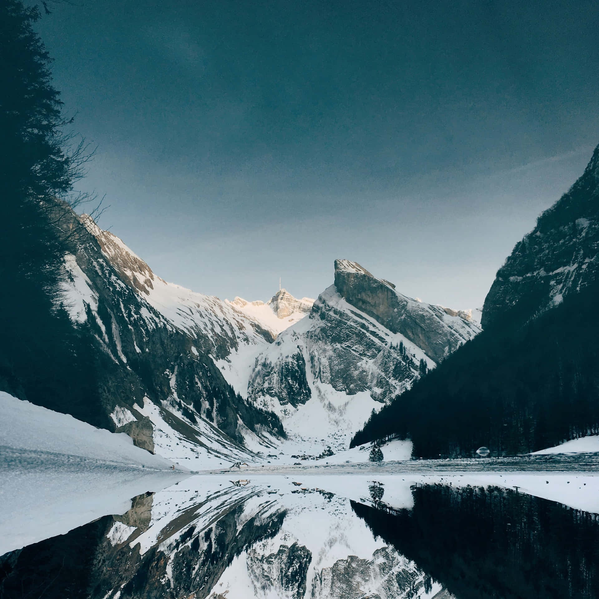  壁紙 — 積雪覆蓋的山峰在冰凍的湖泊後方戲劇性地聳立，湖面映照出它們陡峭的山巔，背景是陰沉的暮色天空。