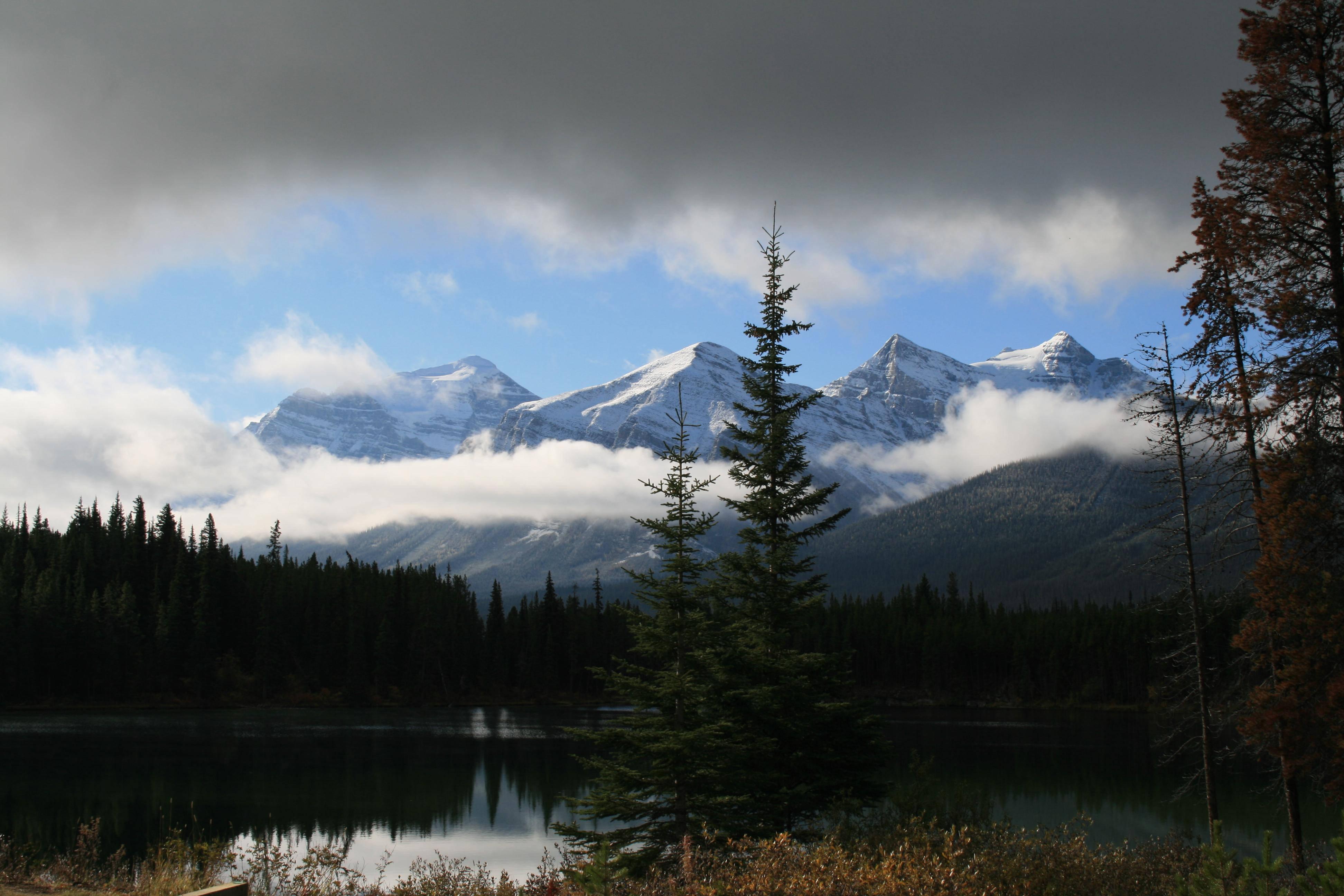 山景 桌布 壁紙 — 積雪覆蓋的山脈在平靜的湖泊後方升起，湖面倒映著雲朵和常綠樹，天空部分多雲。