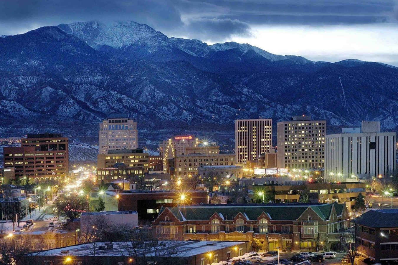 Colorado 桌布 壁紙 — 積雪覆蓋的山脈聳立在黃昏的城市景觀之上，街燈和建築窗戶閃爍著光芒。