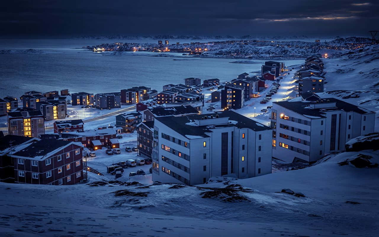 Greenland 桌布 壁紙 — 積雪覆蓋的建築物在俯瞰冰封海洋的黑暗冬日天空中溫暖地發光。