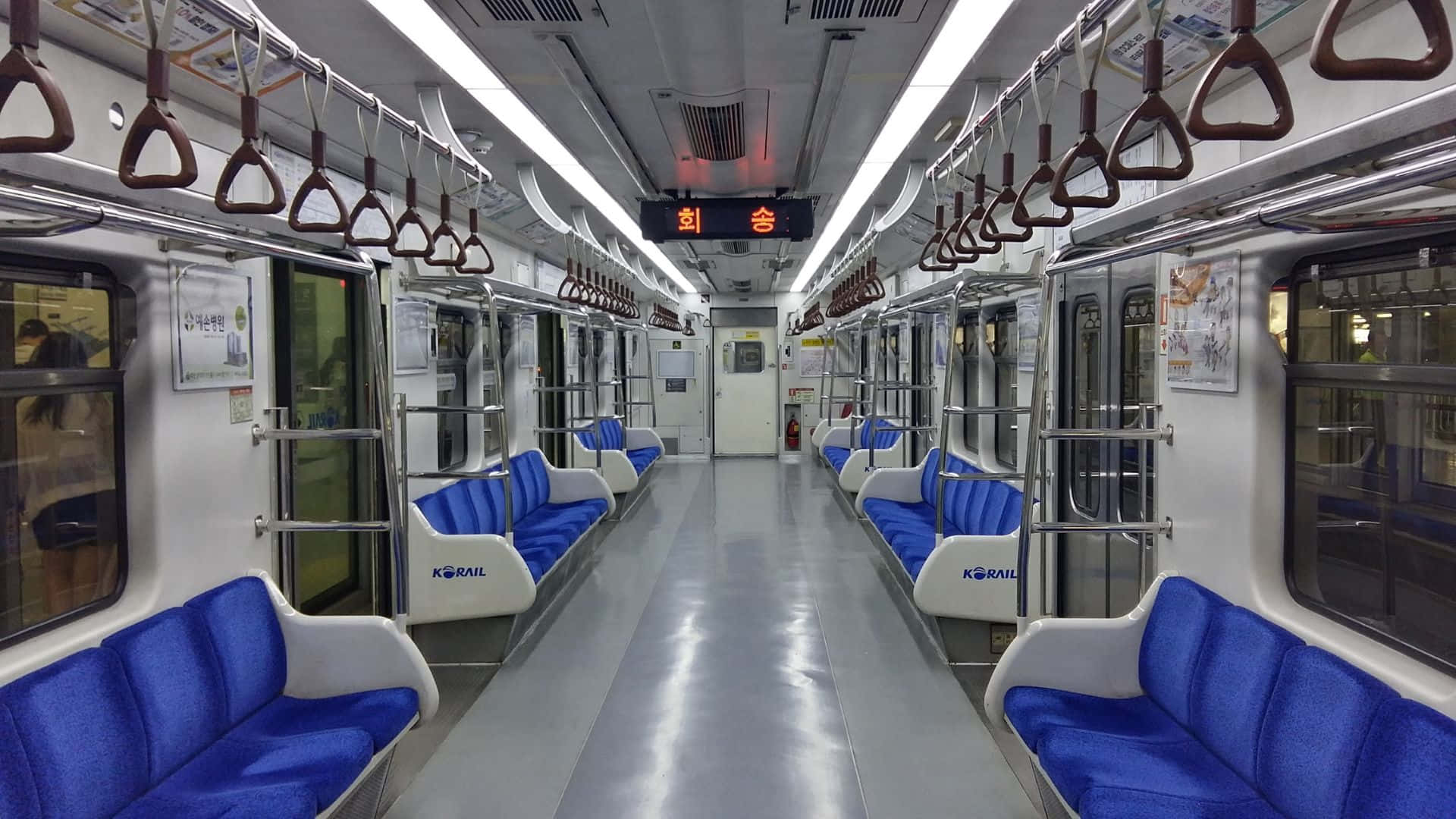 Subway 桌布 壁紙 — 空曠的韓國火車車廂，藍色座椅和頭頂扶手，在明亮的螢光燈下。
