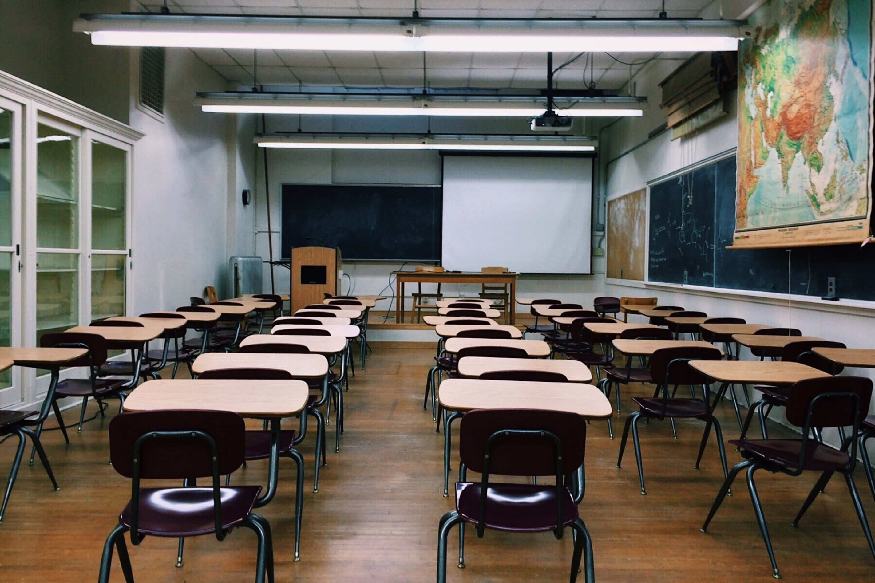 School Photo 桌布 壁紙 — 空蕩蕩的教室，螢光燈下排列著木製書桌和椅子，牆上掛著一個大地圖。