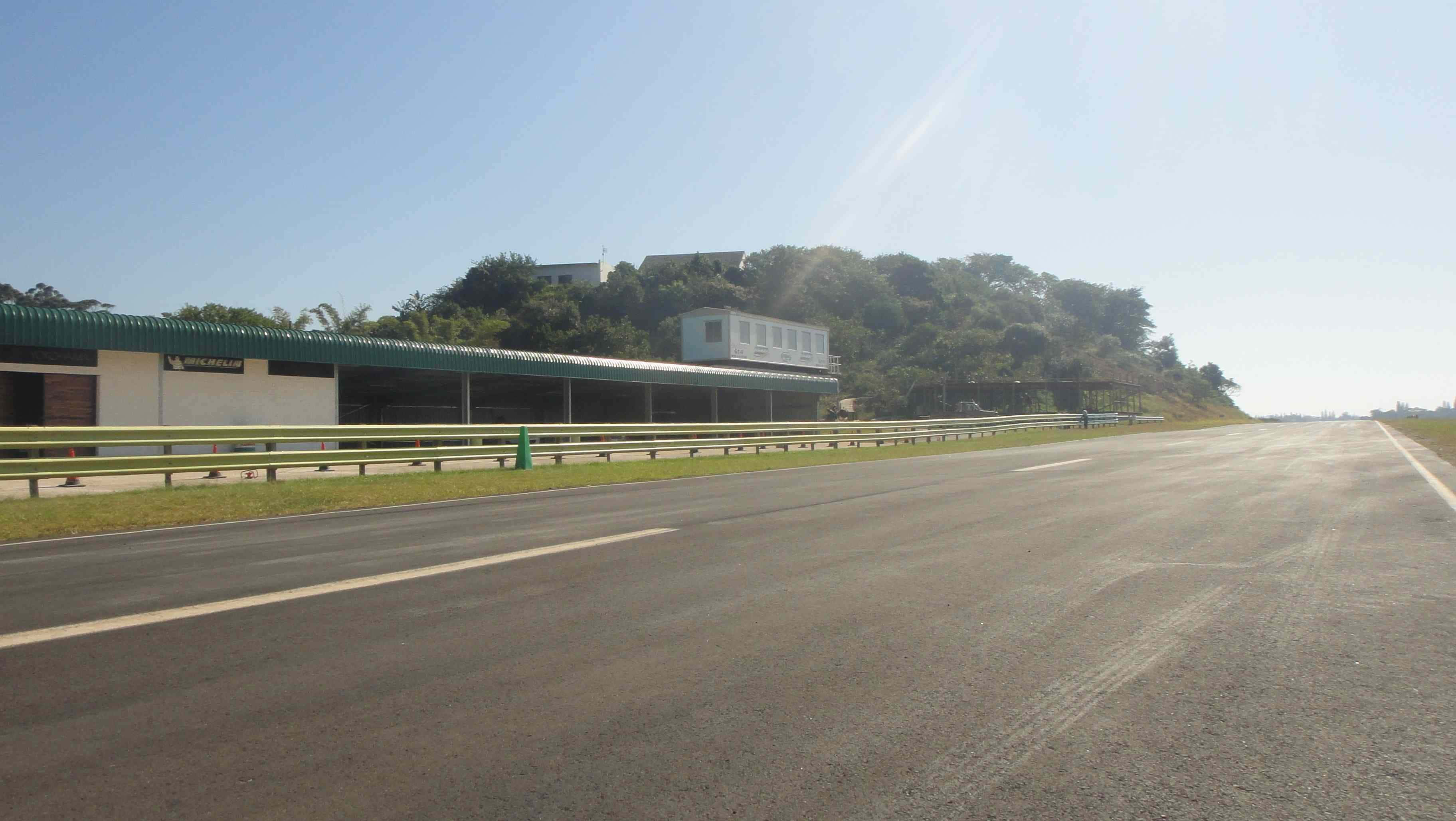 Race Track 桌布 壁紙 — 空蕩蕩的瀝青高速公路在明亮的陽光下延伸，設有護欄和綠色屋頂的建築物，旁邊是樹木覆蓋的山丘。