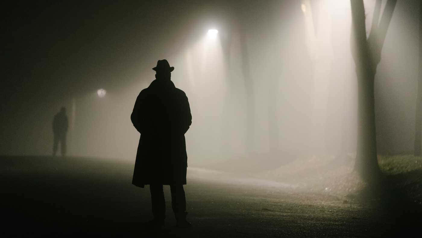 Film Noir 桌布 壁紙 — 穿著 trench coat 和 fedora 帽的剪影人物獨自站在霧氣瀰漫的夜間公園，被投射出詭異光束的路燈照亮。