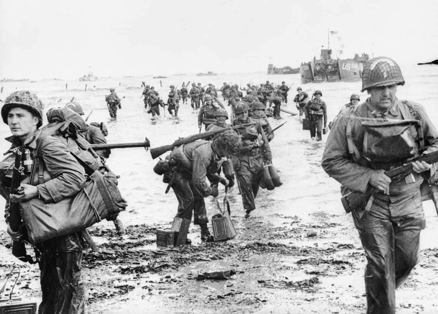 Ww2 桌布 壁紙 — 穿著二戰裝備的士兵在泥濘的海灘上涉水而過，背景有登陸艇。