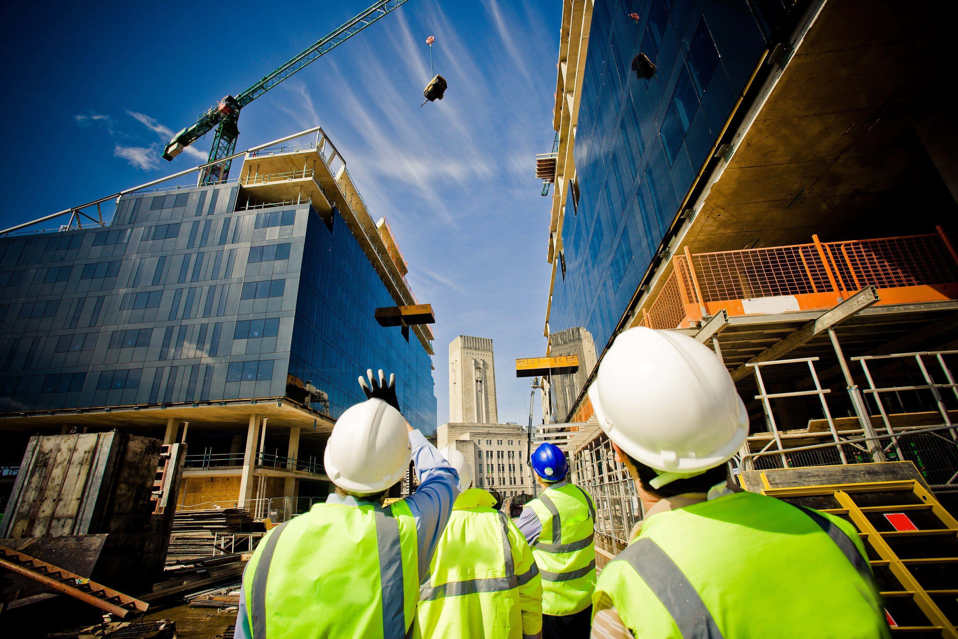 Infrastructure 桌布 壁紙 — 穿著亮黃色背心和白色安全帽的建築工人站在繁忙的城市工地，在晴朗的藍天下，起重機在玻璃帷幕的摩天大樓之間吊運材料。