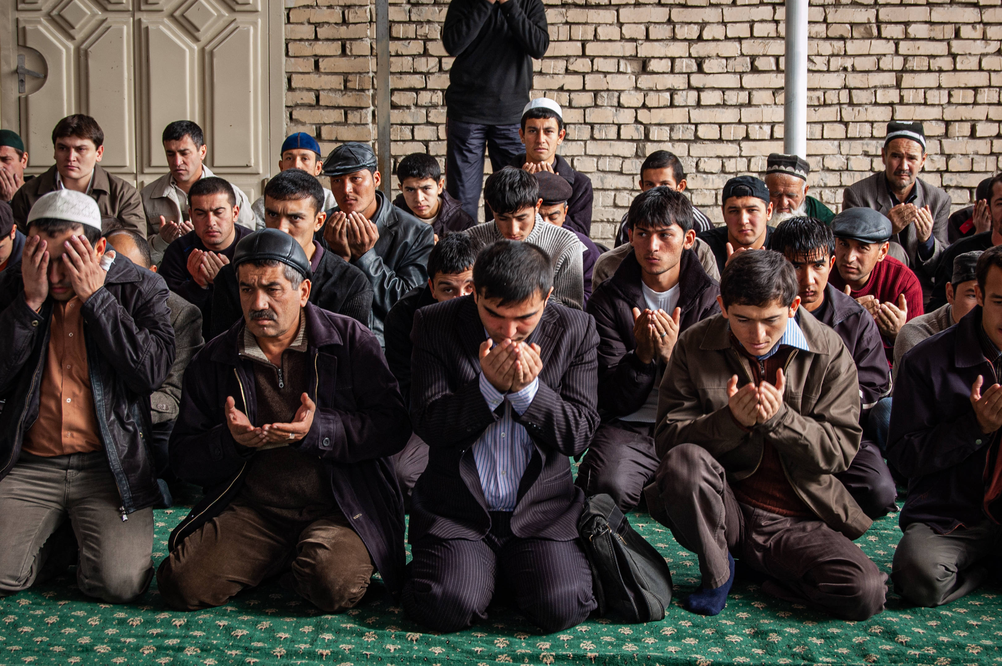 Uzbekistan 桌布 壁紙 — 穿著傳統服飾的男子在磚牆清真寺內的綠色圖案地毯上跪拜祈禱。