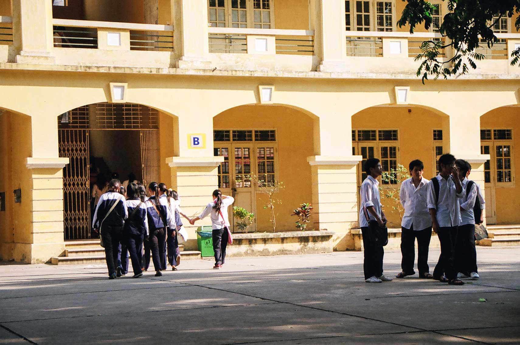 越南 桌布 壁紙 — 穿著制服的學生在陽光普照的黃色殖民風格學校建築外走動和聚集，建築有拱形門口。