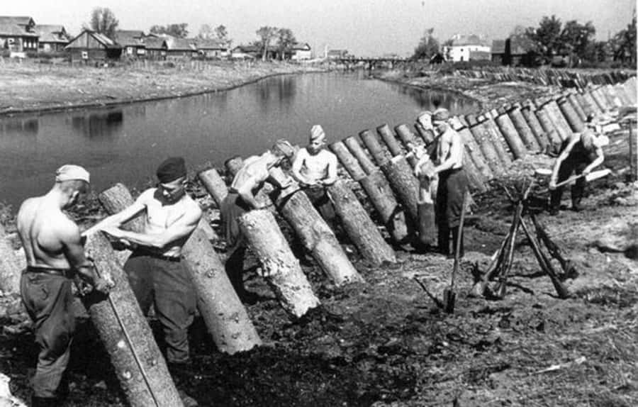 World War 2 桌布 壁紙 — 穿著制服的男人們在鄉村環境中沿著河岸辛勤地堆放巨大的原木。