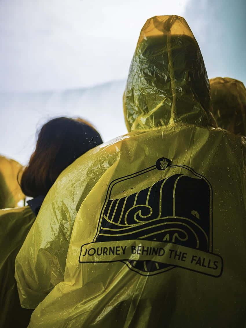 Yellow Raincoat 桌布 壁紙 — 穿著印有「Journey Behind the Falls」標誌的黃色雨衣斗篷的人，背對鏡頭，站在霧氣瀰漫的瀑布附近。