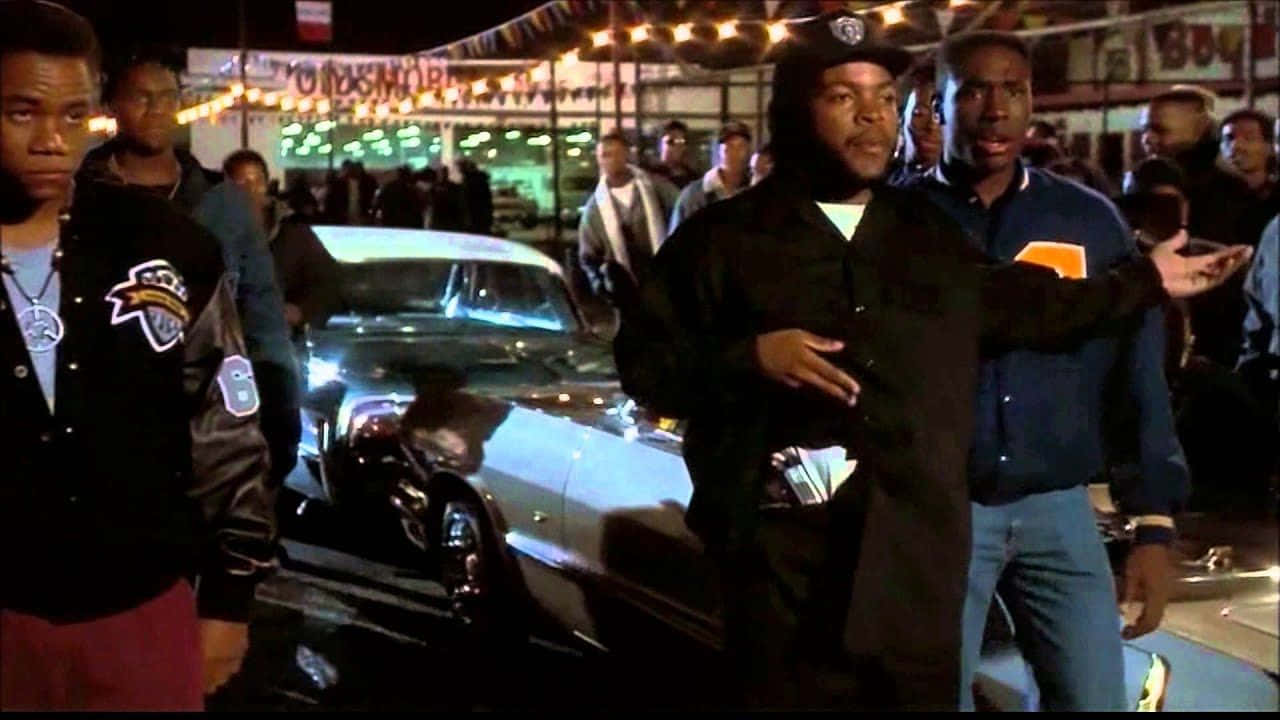 Boyz N The Hood 桌布 壁紙 — 穿著外套的男人站在閃亮的經典汽車周圍，在夜間戶外市場的燈串下。