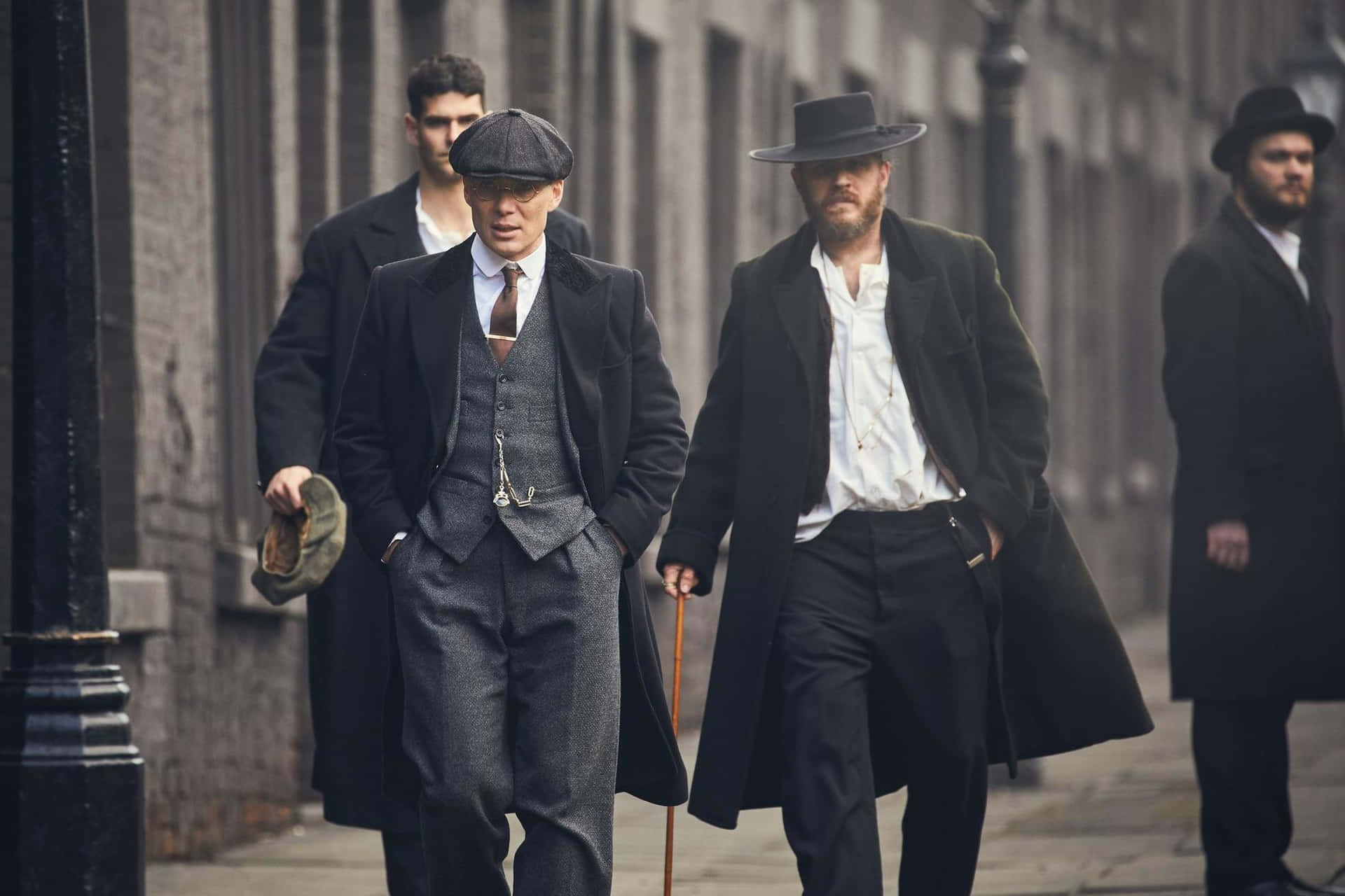 四個穿著復古大衣和帽子的男人走在城市街道上。 — Peaky Blinders 壁紙