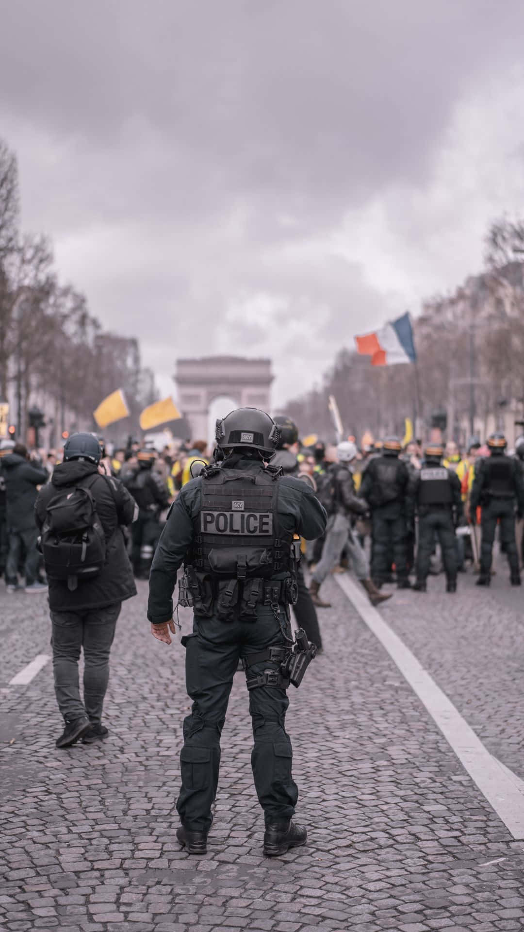 cop 桌布 壁紙 — 穿著戰術裝備的警察站在鵝卵石街道上，面對凱旋門附近的人群，天空多雲。