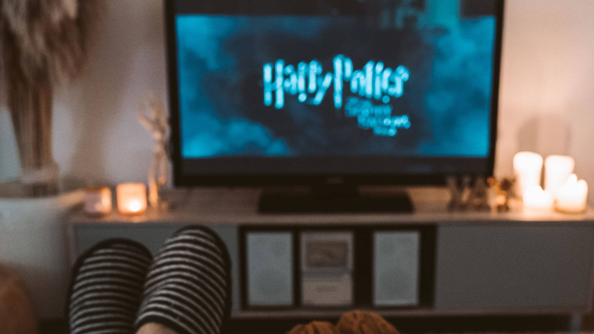 watching tv 桌布 壁紙 — 穿著條紋襪子的舒適雙腳在電視前休息，電視正在播放哈利波特，媒體櫃上閃爍著溫暖的燭光。