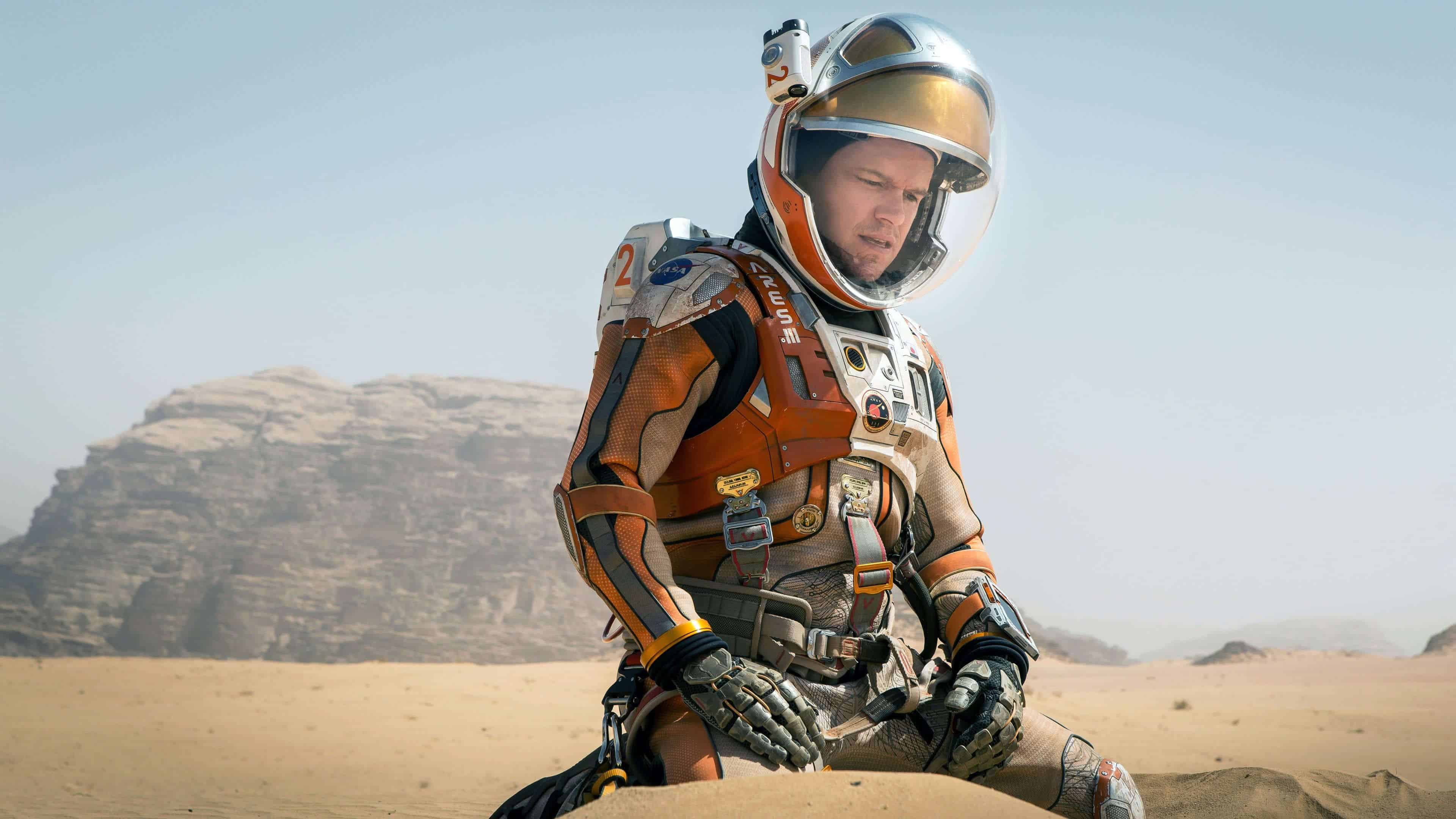 Matt Damon 桌布 壁紙 — 穿著橙色和白色太空衣的太空人跪在荒蕪的沙漠沙地上，背景有岩石峭壁，在明亮的日光下。