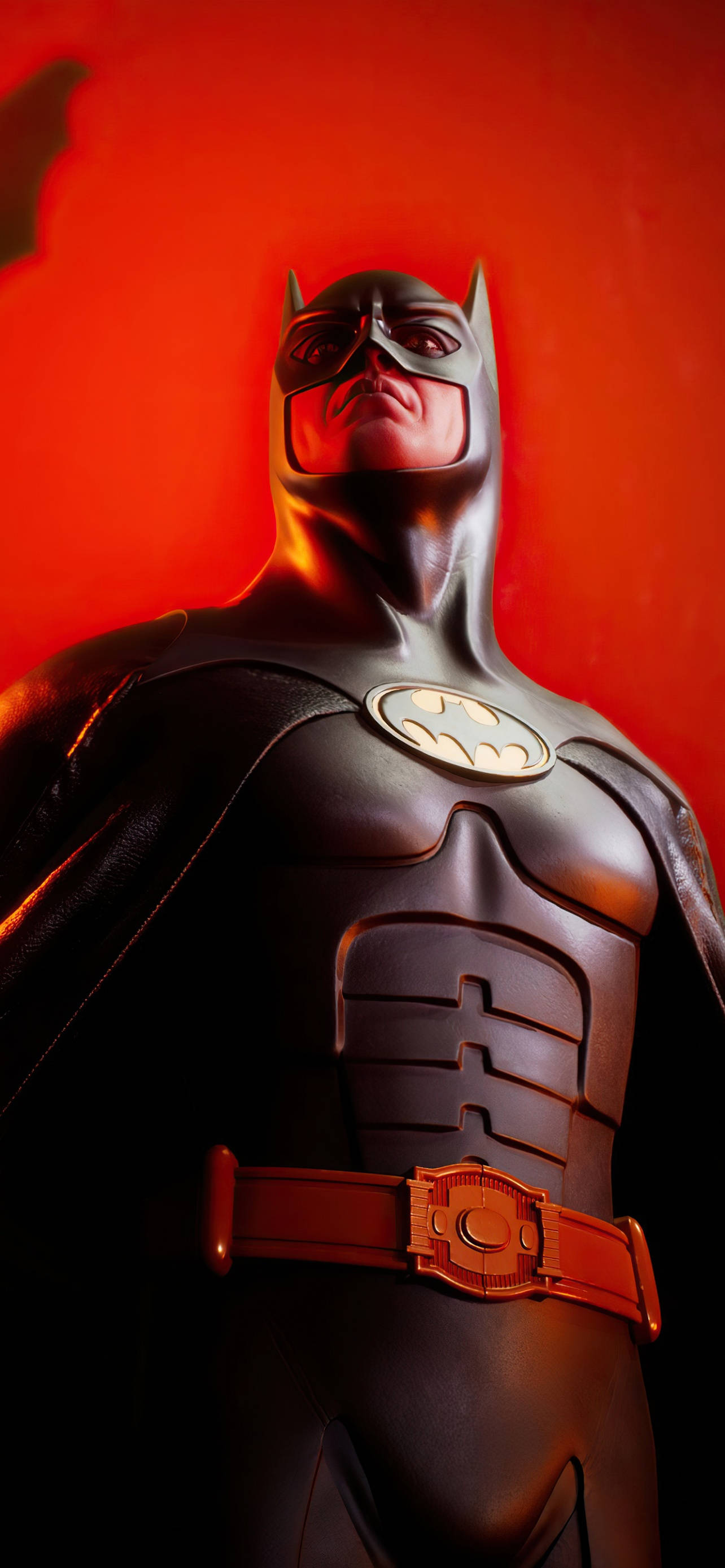 The Batman iPhone 桌布 壁紙 — 穿著深色西裝的蝙蝠俠雕像站在發光的紅色背景前，戲劇性的燈光凸顯了他裝甲的胸膛和披風。