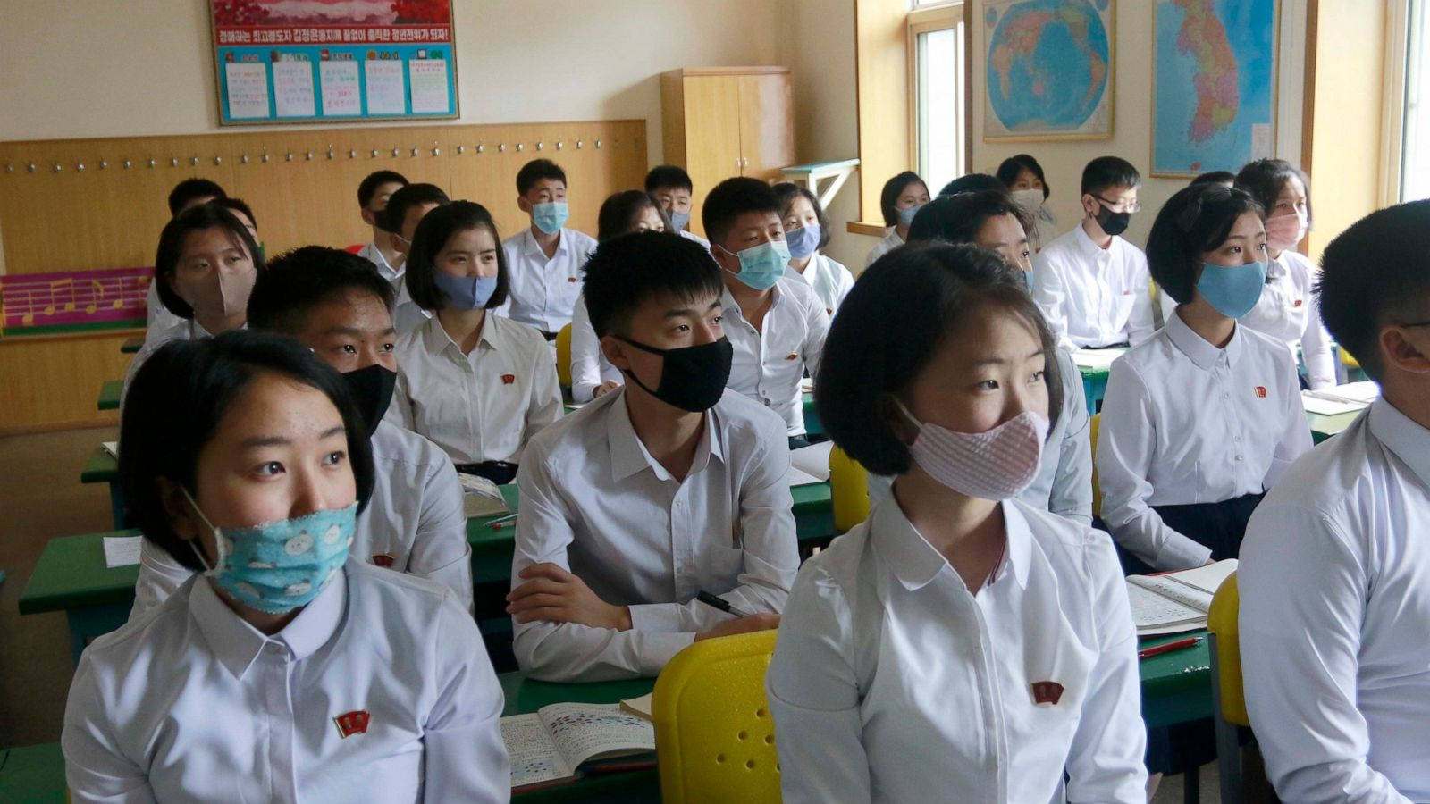 Coronavirus 桌布 壁紙 — 穿著白色制服和口罩的學生們在明亮的教室裡專心坐著，牆上掛著地圖和海報。