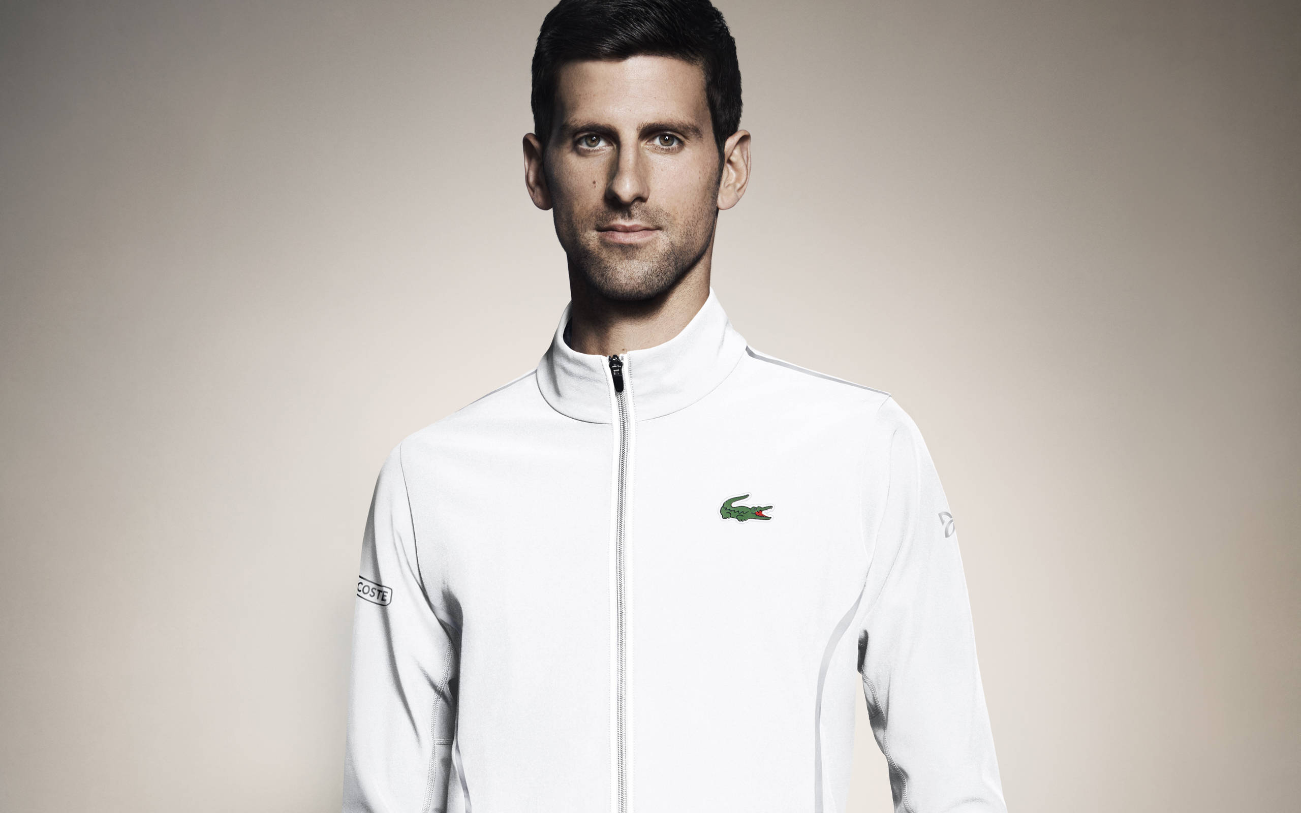 Novak 桌布 壁紙 — 穿著白色拉鍊外套、帶有綠色鱷魚標誌的認真網球運動員，在中性工作室背景下直視鏡頭。