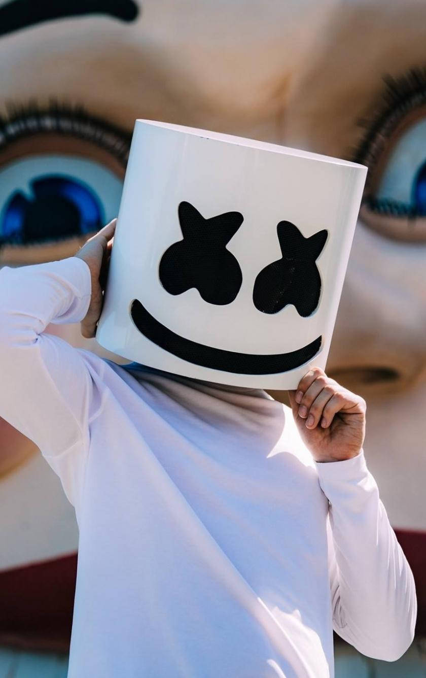 marshmello iPhone 桌布 壁紙 — 穿著白色襯衫的人手持標誌性的白色棉花糖面具，上面有黑色笑臉，背景是巨大的娃娃眼睛。