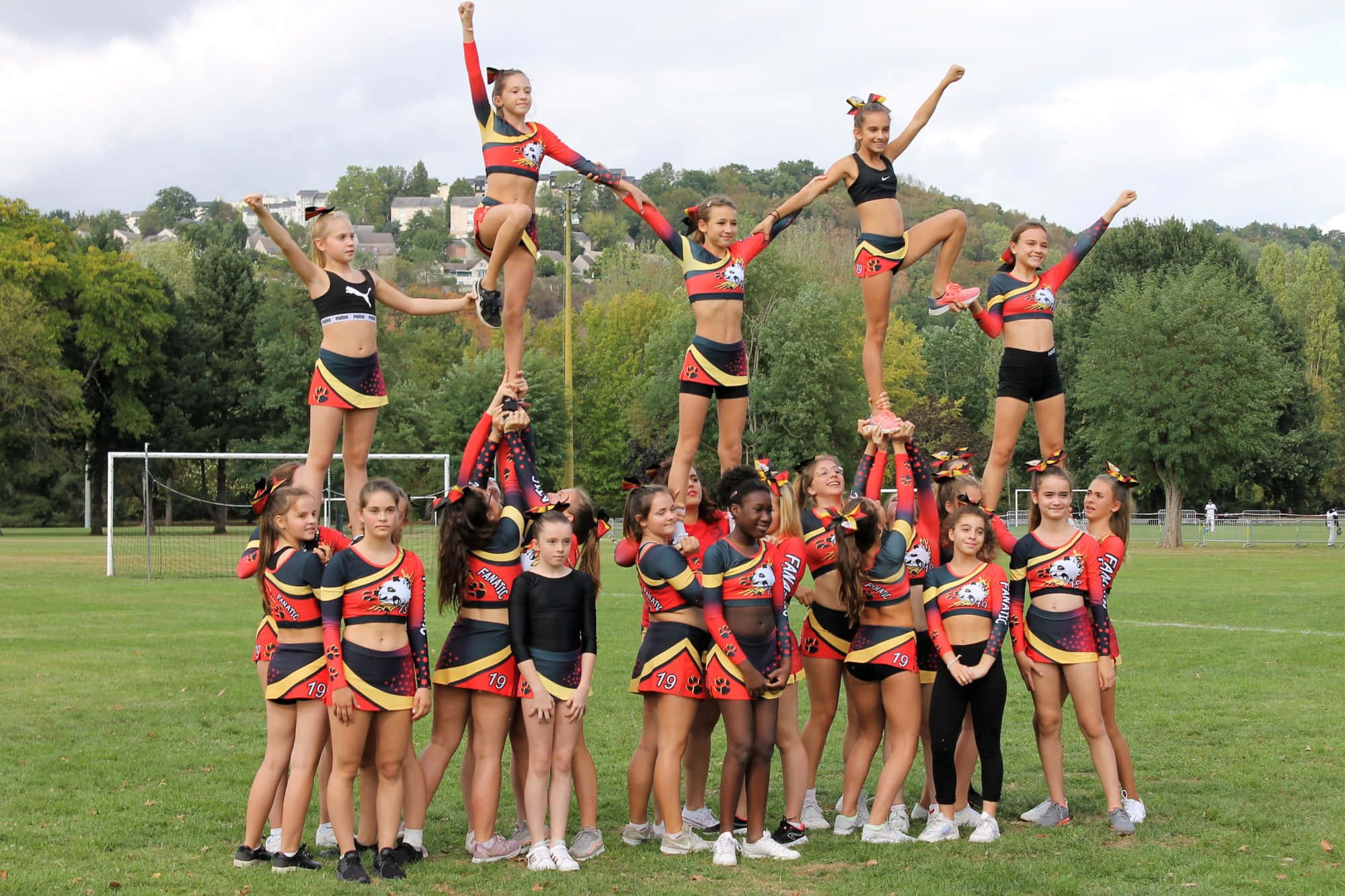 Cheer 桌布 壁紙 — 穿著紅、黑、黃色制服的啦啦隊在草地上以金字塔陣式擺出動感姿勢，背景有樹木和房屋。