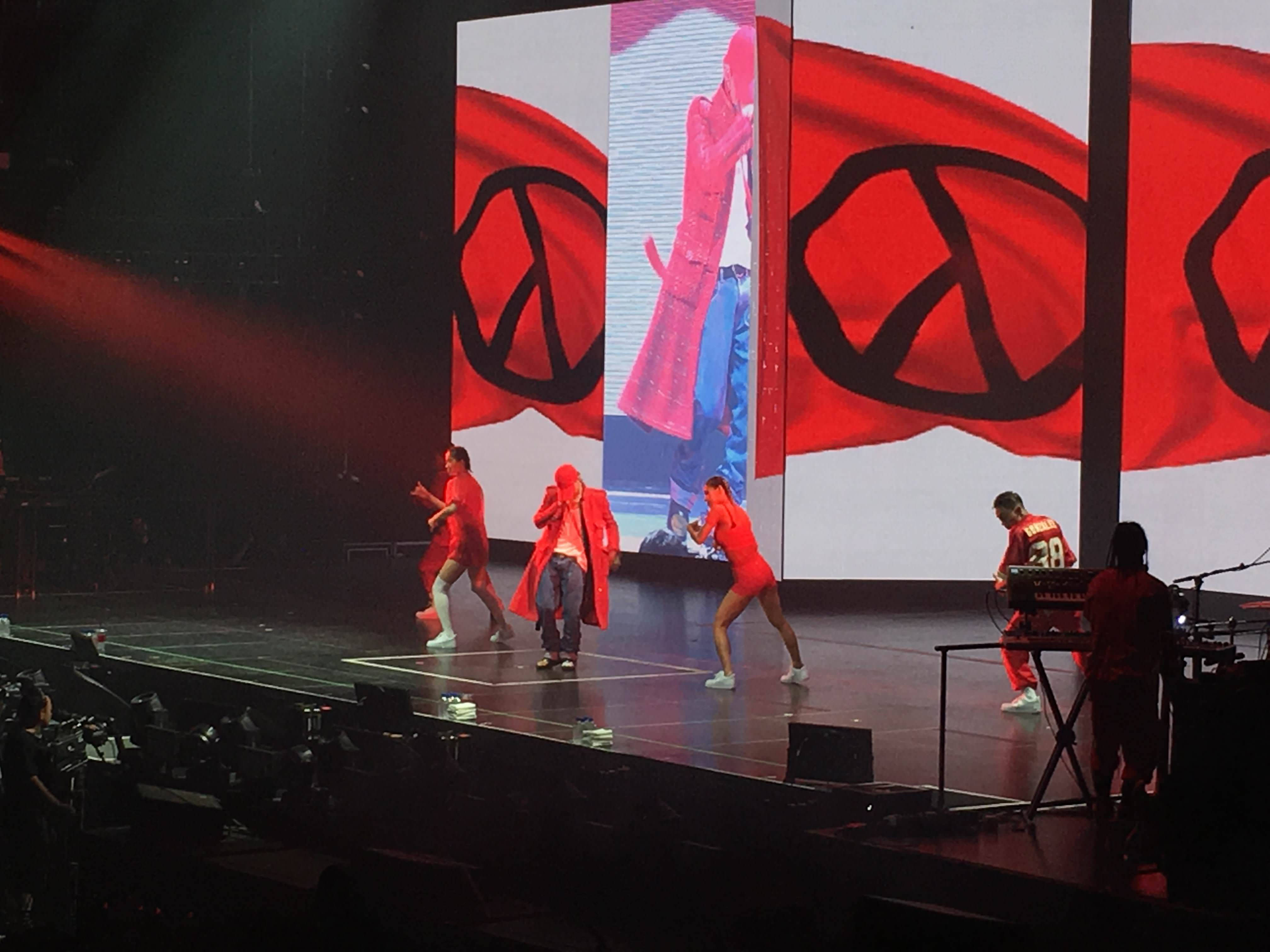 G-dragon 桌布 壁紙 — 穿著紅色服裝的表演者在昏暗的舞台上，在紅色聚光燈下，背後有大型和平符號螢幕，充滿活力地跳舞。