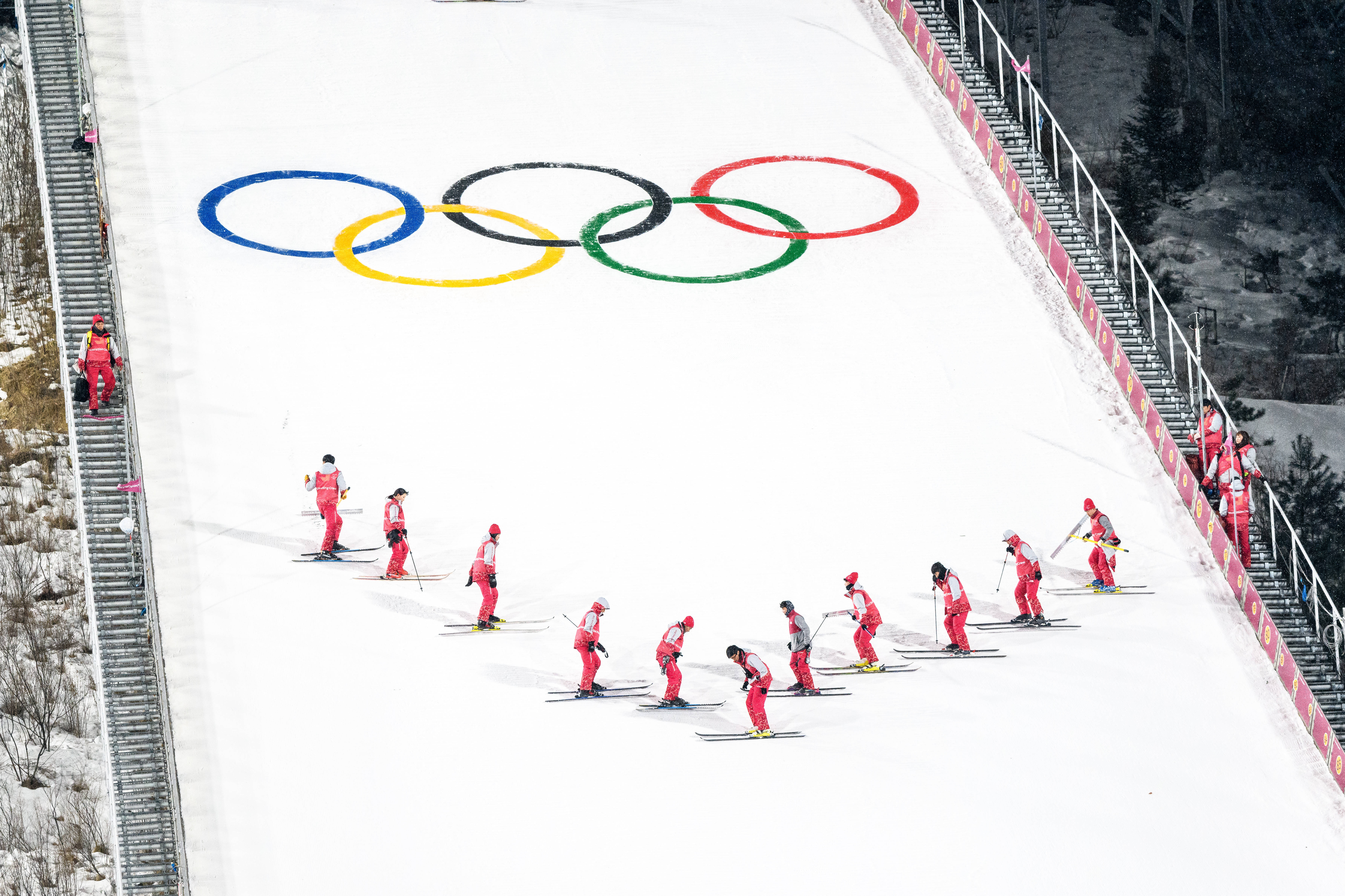 Olympics 桌布 壁紙 — 穿著紅色滑雪服的滑雪者在雪地上畫有巨型奧運五環的雪坡上排隊。