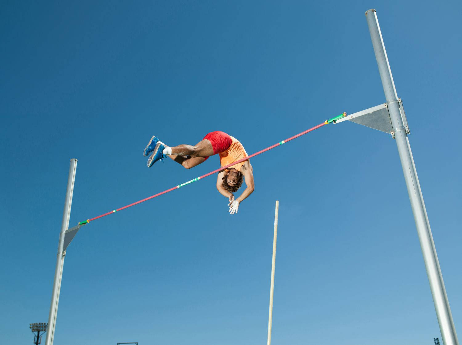 Athletic 桌布 壁紙 — 穿著紅色短褲和橙色襯衫的運動員在晴朗的藍天下完美地執行了福斯貝利式跳高。