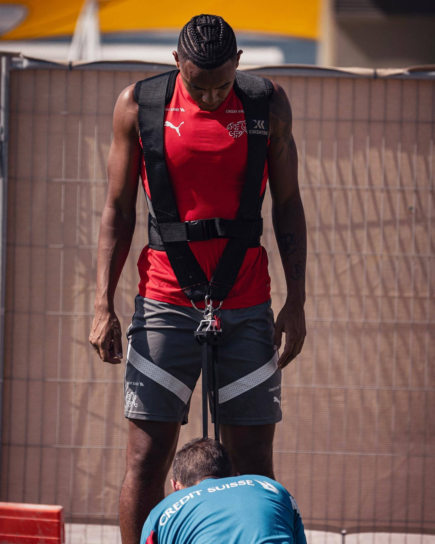 Football Players 桌布 壁紙 — 穿著紅色訓練裝備的運動員在明亮陽光下高高站立，而教練在下方調整安全帶。
