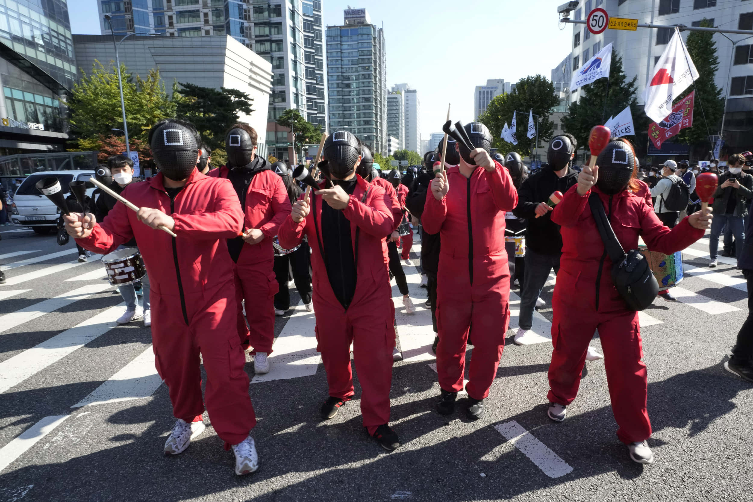 Halloween Parades 桌布 壁紙 — 穿著紅色連身衣和黑色面具的人們手持沙鈴和鼓在城市的街道上游行。